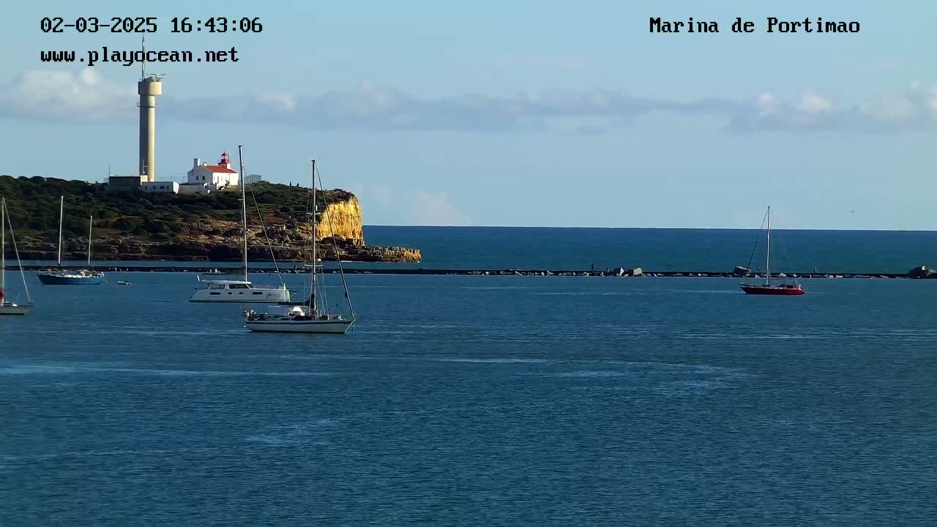Several sailboats are moored in a calm harbor near a rocky cliff with a lighthouse and a tall tower under a mostly clear sky.
