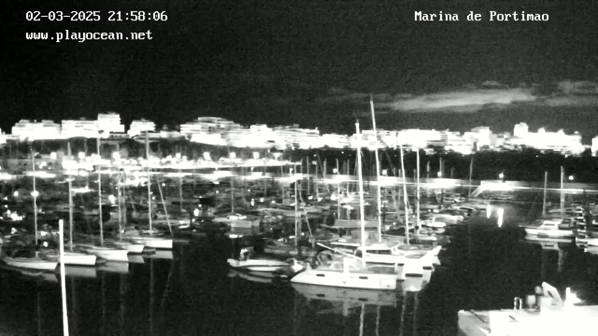 A nighttime view of a marina filled with numerous boats, with a city illuminated in the background under a partly cloudy sky.