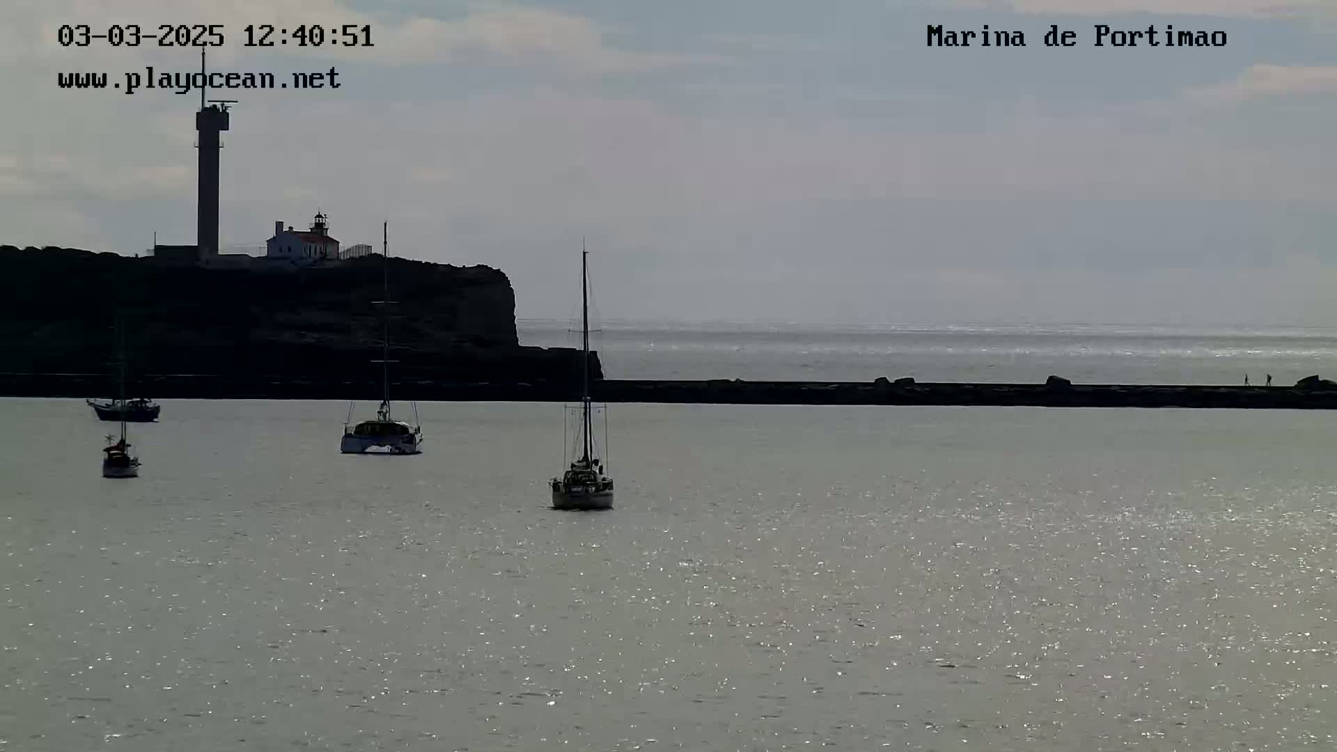 Several sailboats are anchored in a calm harbor near a lighthouse on a partly cloudy day.