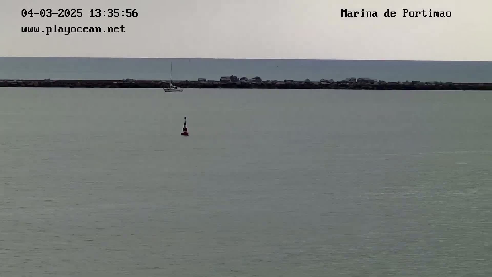 A calm, gray day shows a mostly still body of water with a small sailboat and a buoy, behind a low rock wall.