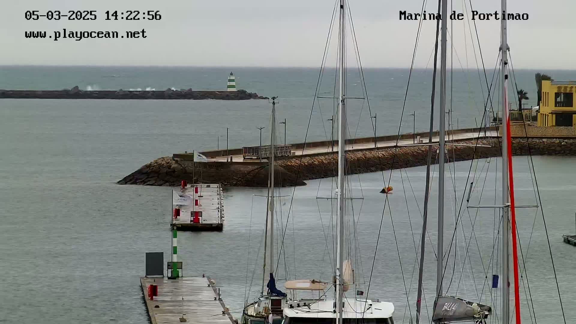 A marina with several sailboats moored, a breakwater with waves crashing against it, and a lighthouse in the distance on an overcast day.