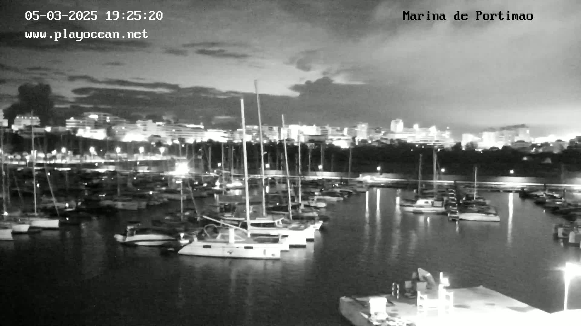 A nighttime view of a marina filled with sailboats and yachts under a partly cloudy sky.