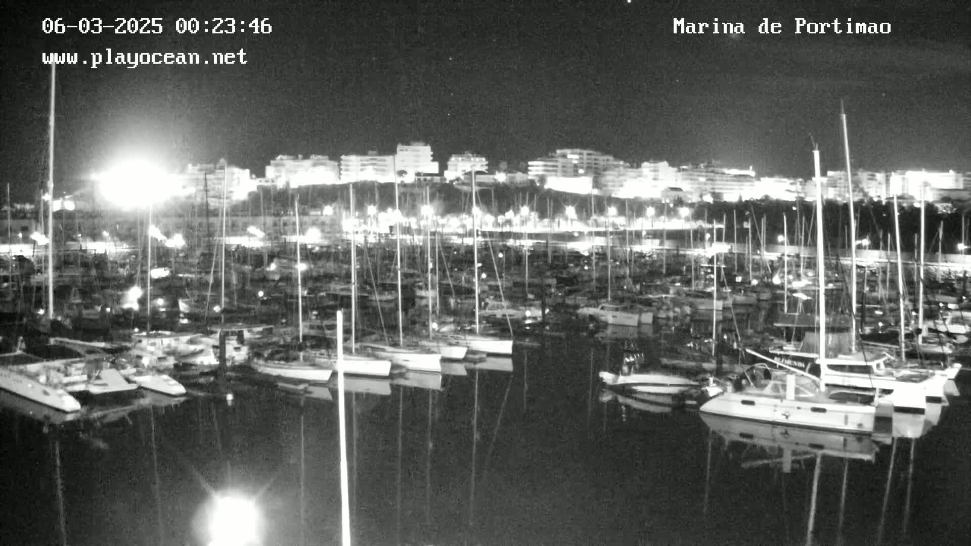A nighttime, black-and-white view of a marina filled with numerous sailboats and motorboats, docked along a waterfront with buildings illuminated in the background.
