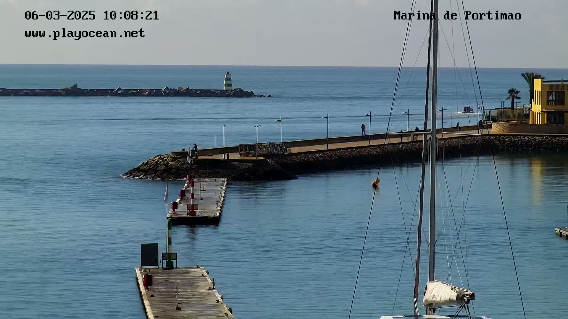 A calm harbor scene on a sunny day shows a marina with several docks, a lighthouse in the distance, and a sailboat in the foreground.