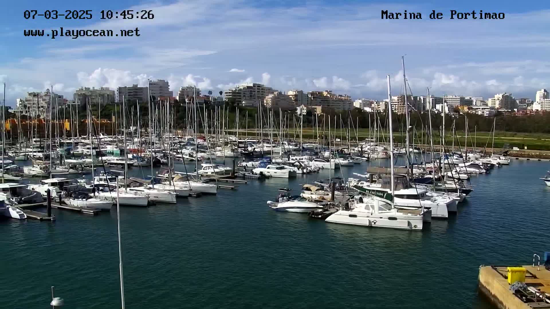 A marina filled with numerous sailboats and motorboats is situated under a partly cloudy sky.