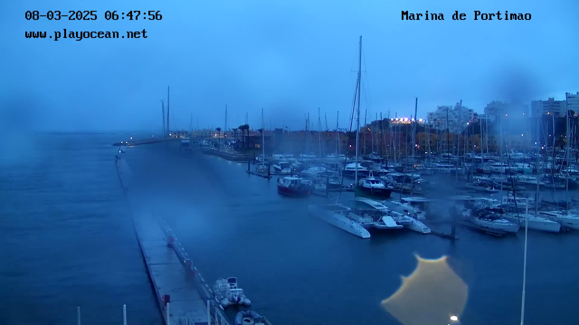 A marina filled with numerous boats is seen on an overcast morning, with rain obscuring the view.