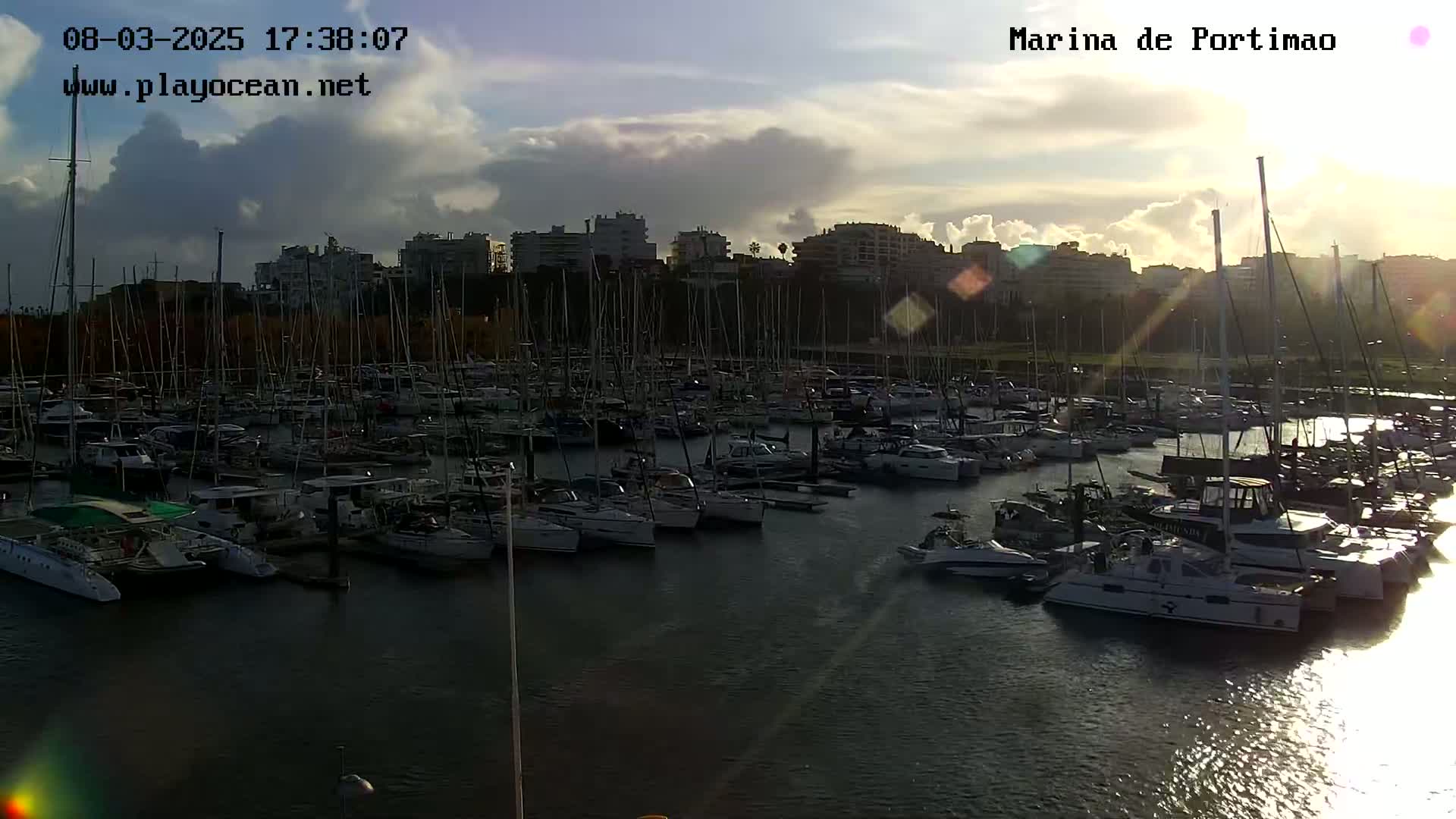 A marina filled with sailboats is seen at sunset under a partly cloudy sky.