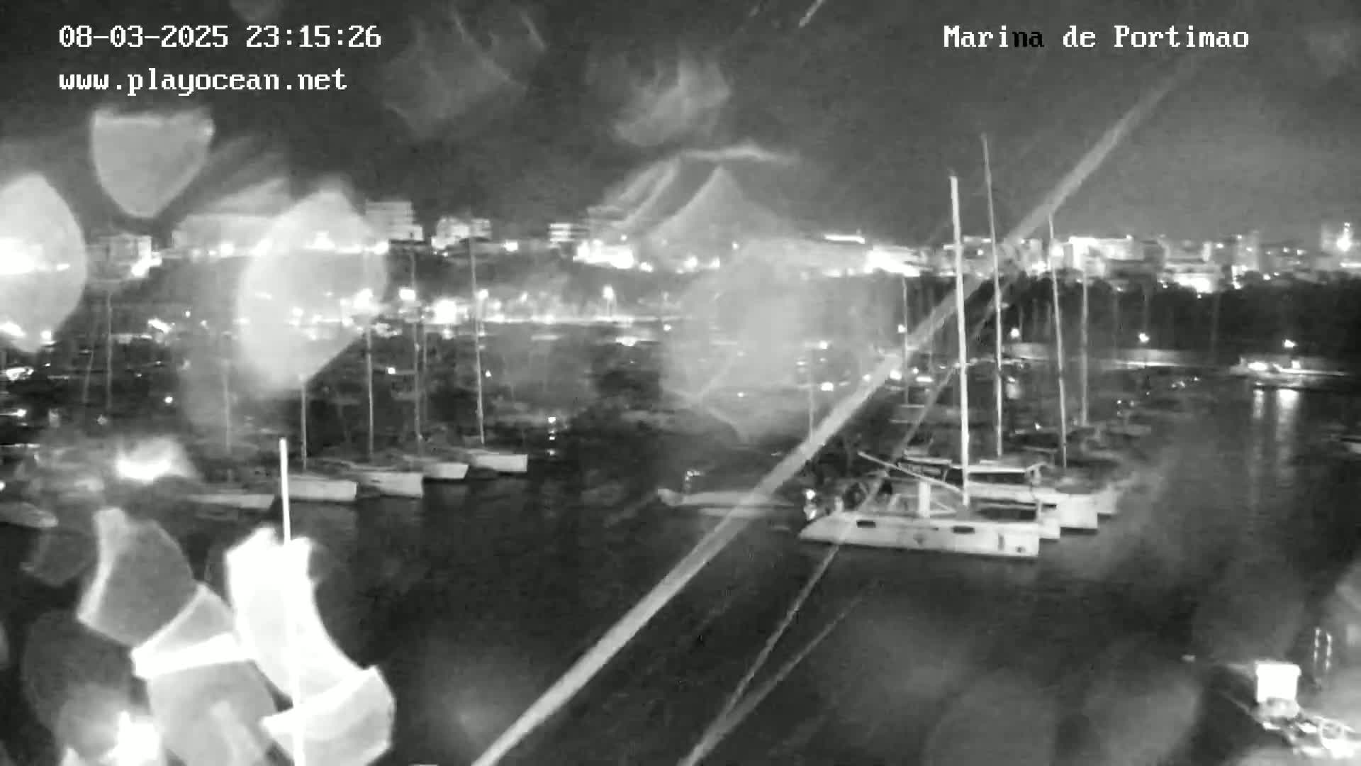 A nighttime, rain-streaked view of a marina filled with sailboats and a city skyline in the background.