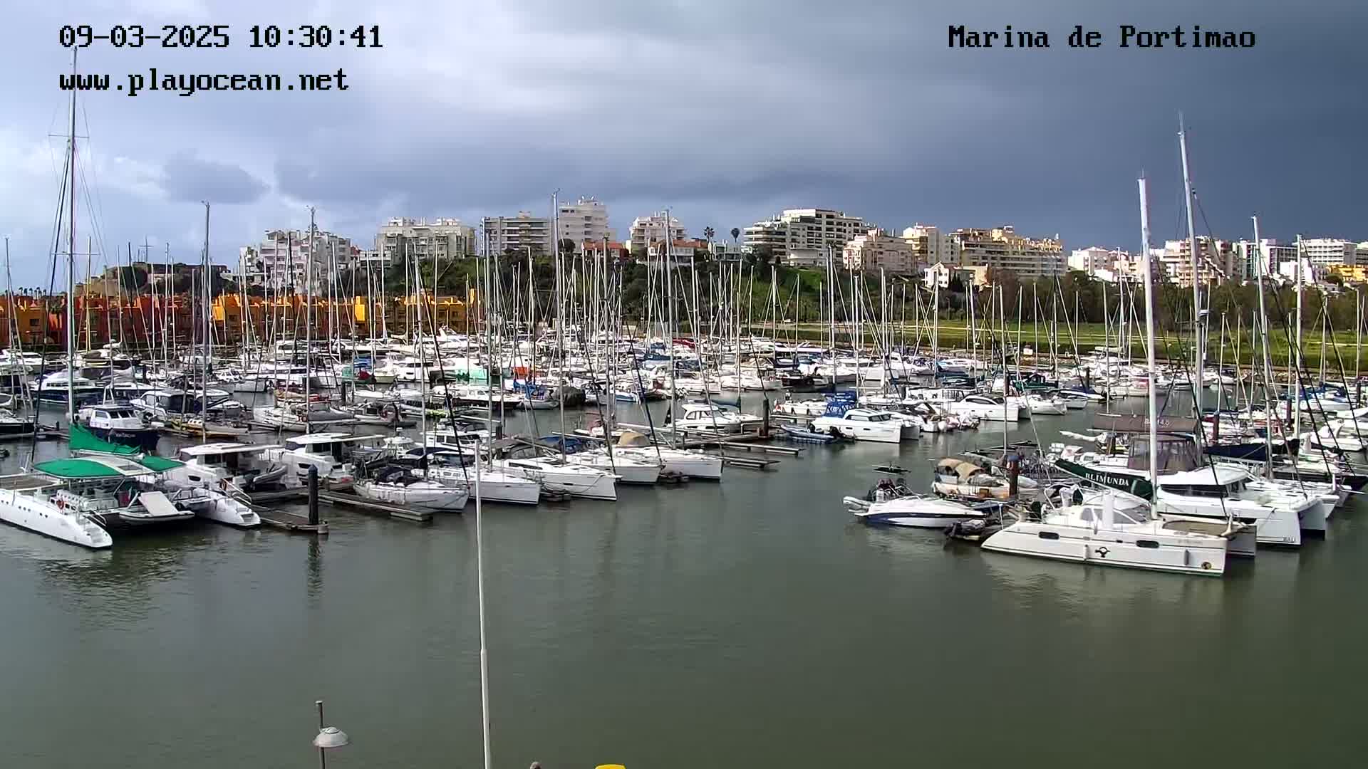 A marina filled with numerous sailboats and motorboats is shown under a partly cloudy sky.