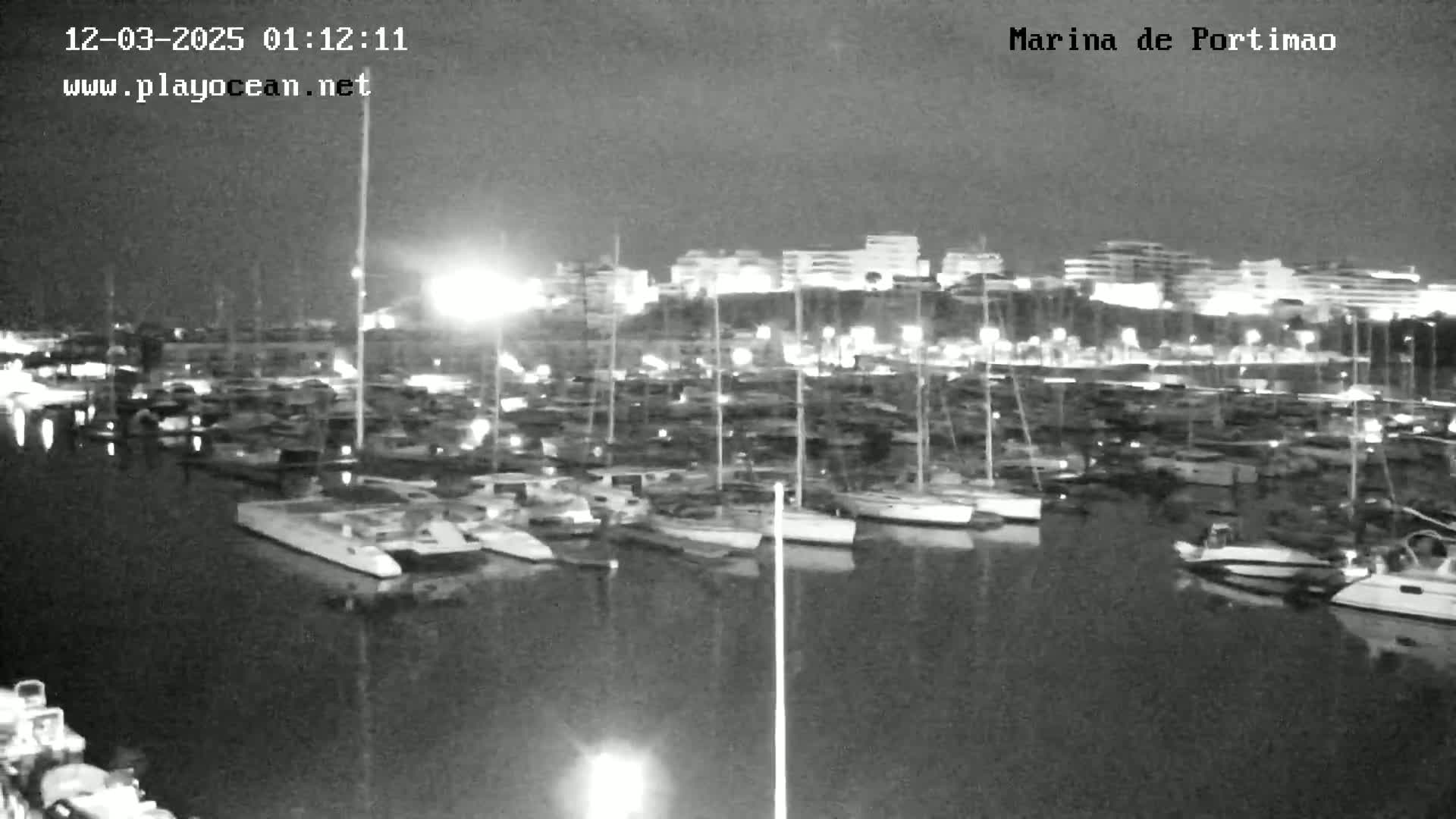 A nighttime, low-light view of a marina filled with numerous boats and yachts docked along a waterfront, with city lights visible in the background.