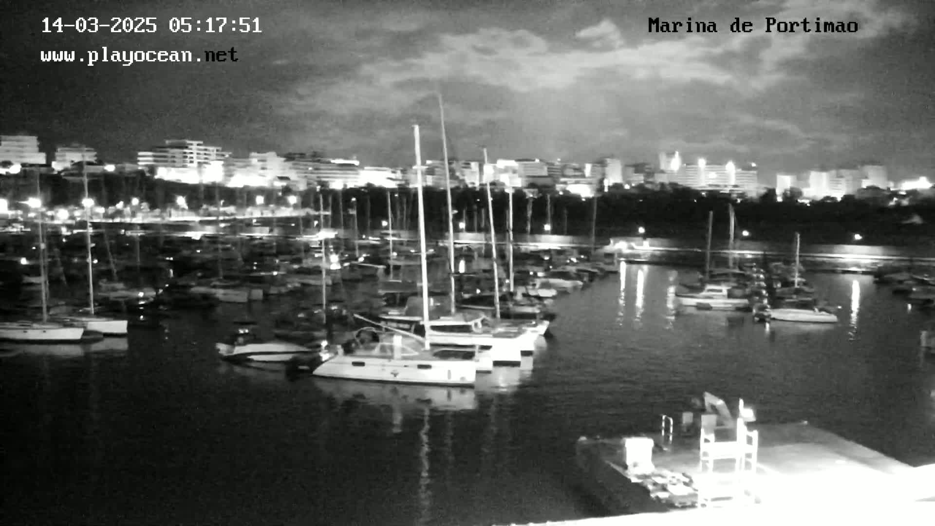A nighttime view of a marina filled with numerous sailboats and yachts, under a partly cloudy sky.
