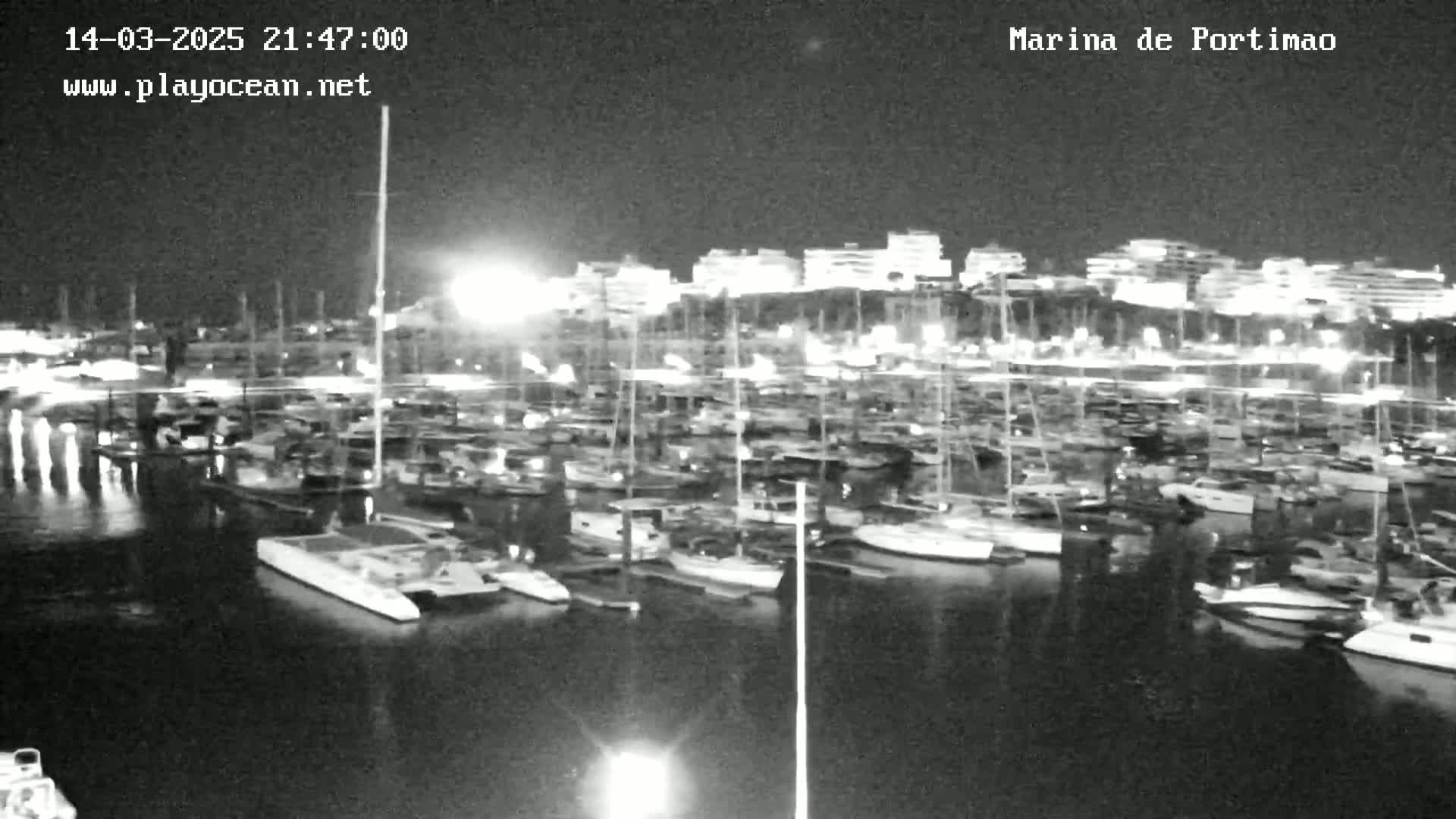 A nighttime, black and white view of a marina filled with numerous boats and yachts, with illuminated buildings visible in the background.