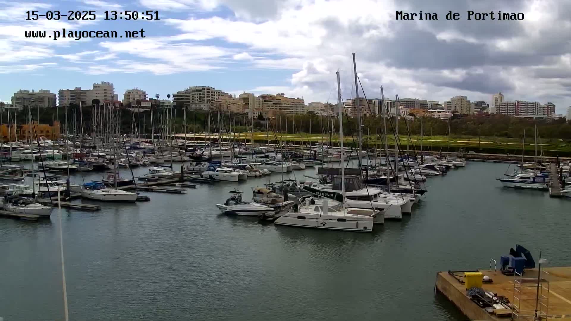 A marina filled with numerous sailboats and motorboats is situated under a partly cloudy sky.