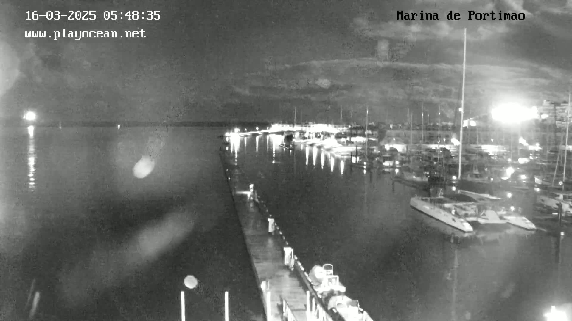 A nighttime, black and white view of a marina filled with boats, calm water, and a pier extending into the water.