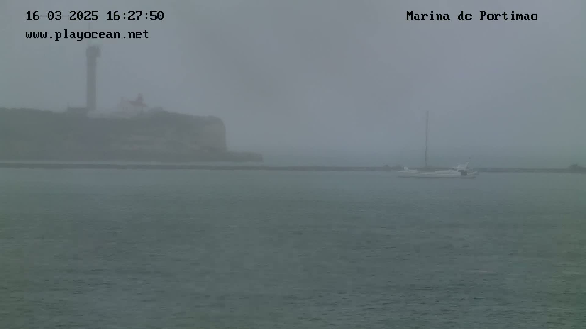 A hazy, foggy view of a calm body of water shows a distant cliff with a tall structure, and a small sailboat at anchor in the foreground.