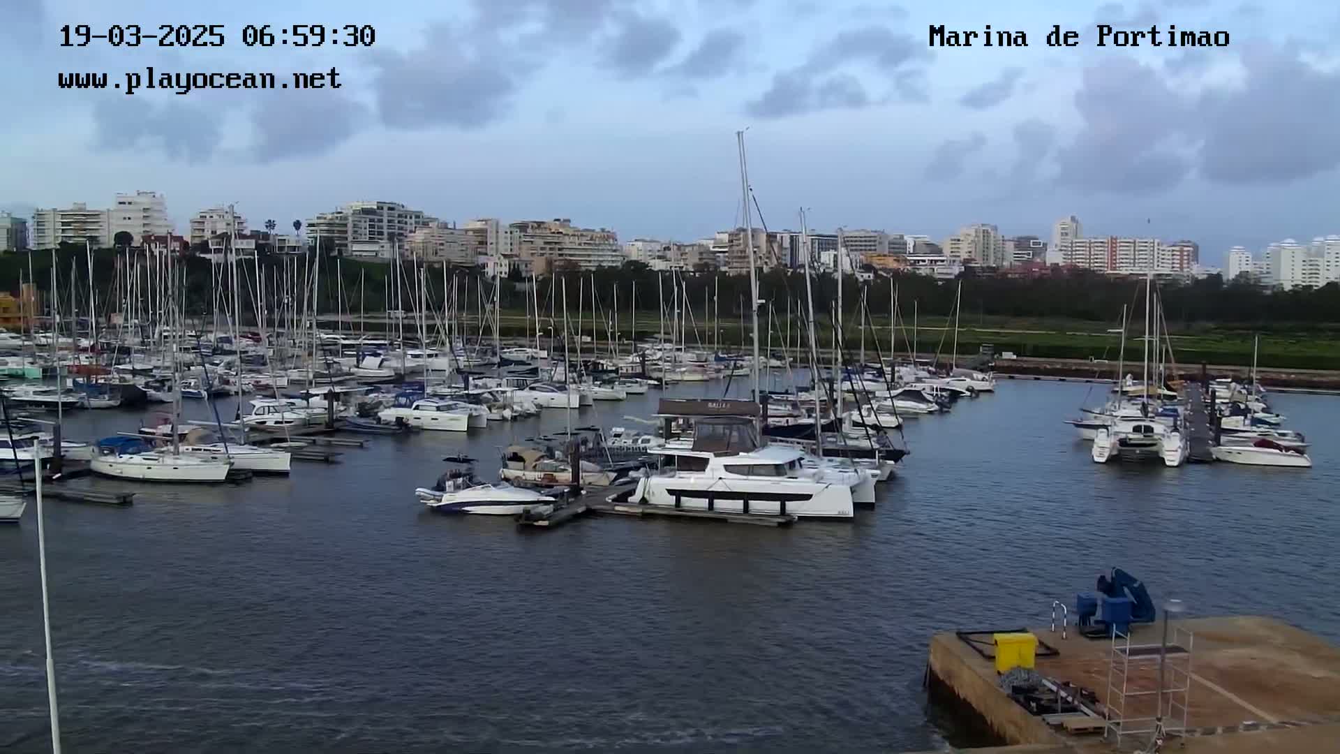 A marina filled with numerous sailboats and motorboats is situated near a city under a partly cloudy sky.