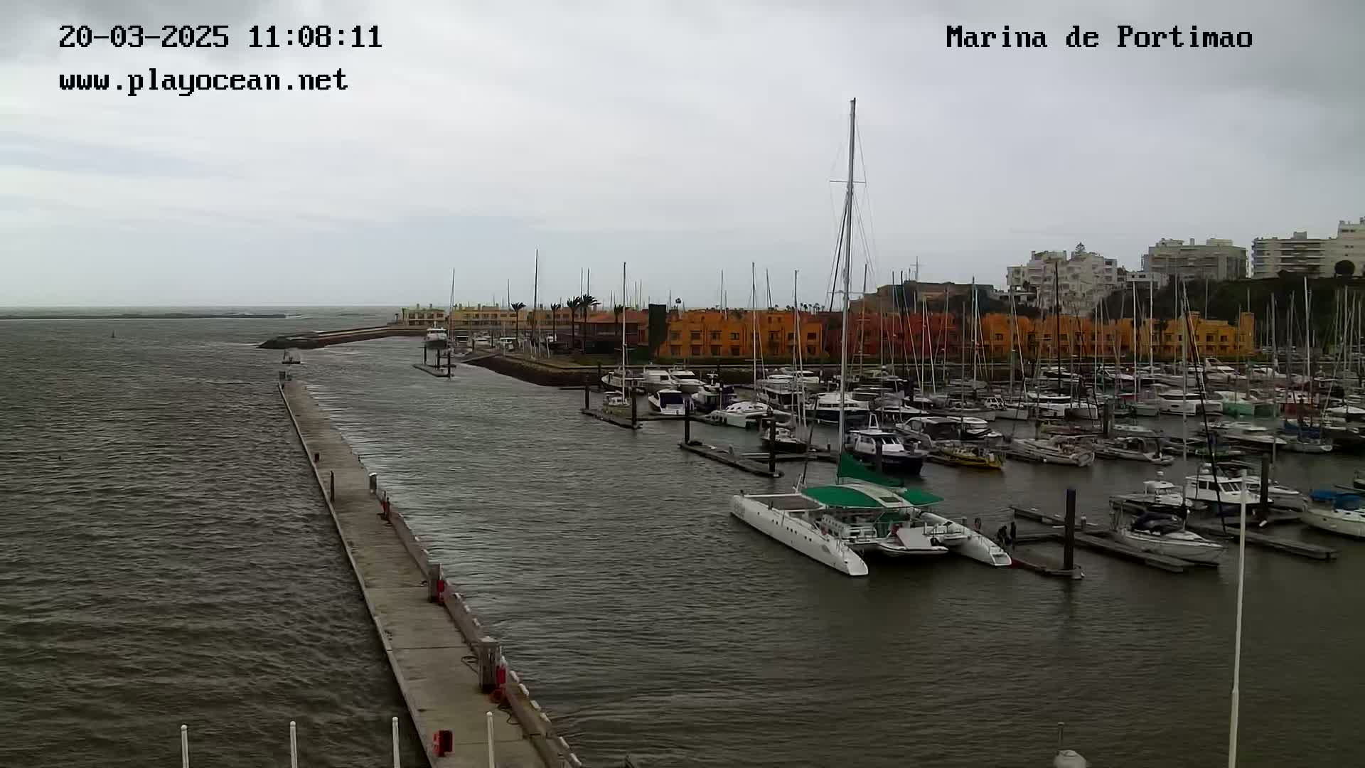A marina filled with numerous boats and yachts is seen on an overcast day, with a long concrete pier extending from the shore into the calm water.