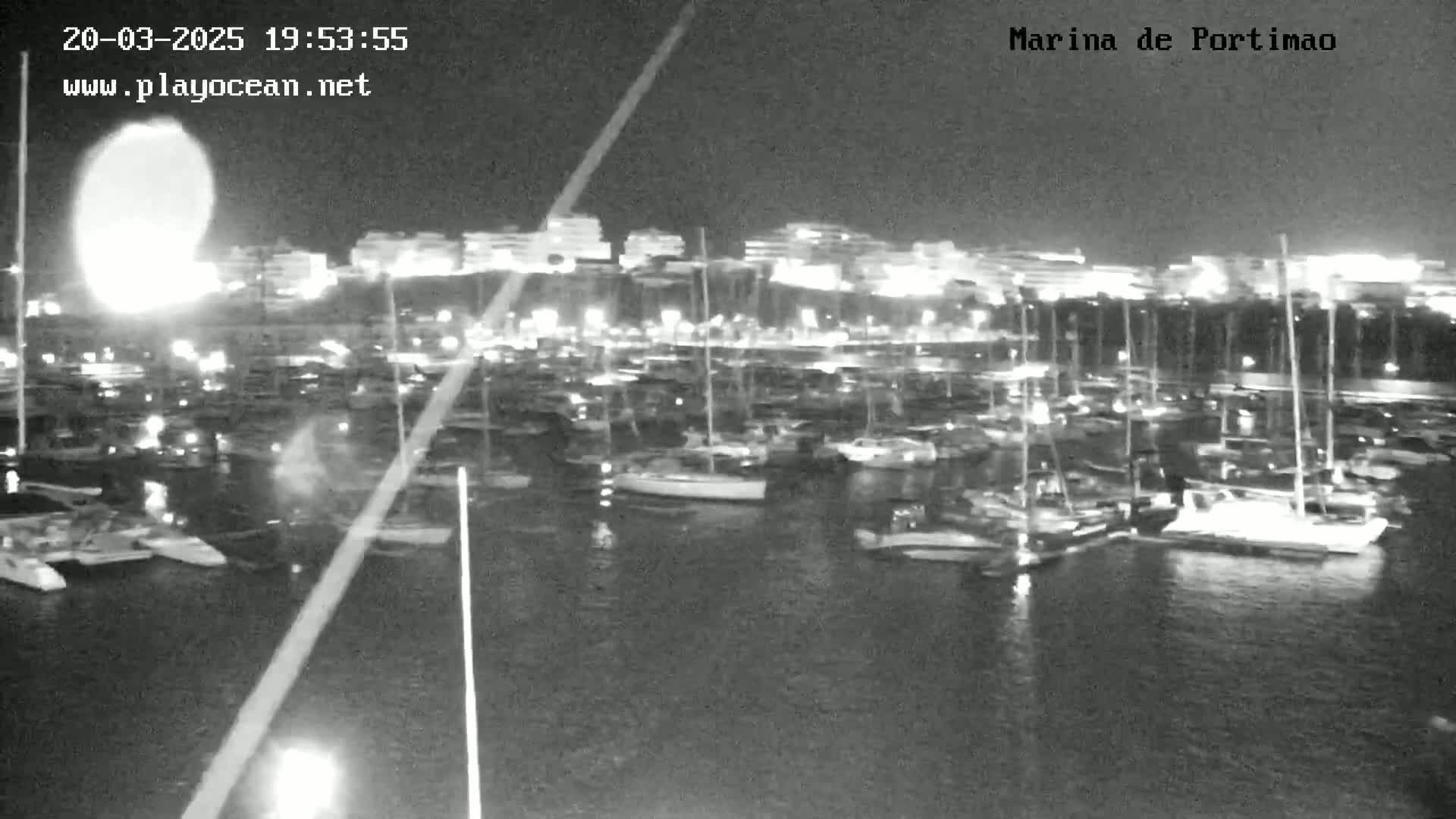 A nighttime, low-light view of a marina filled with numerous boats and yachts, illuminated by the lights of the surrounding buildings.