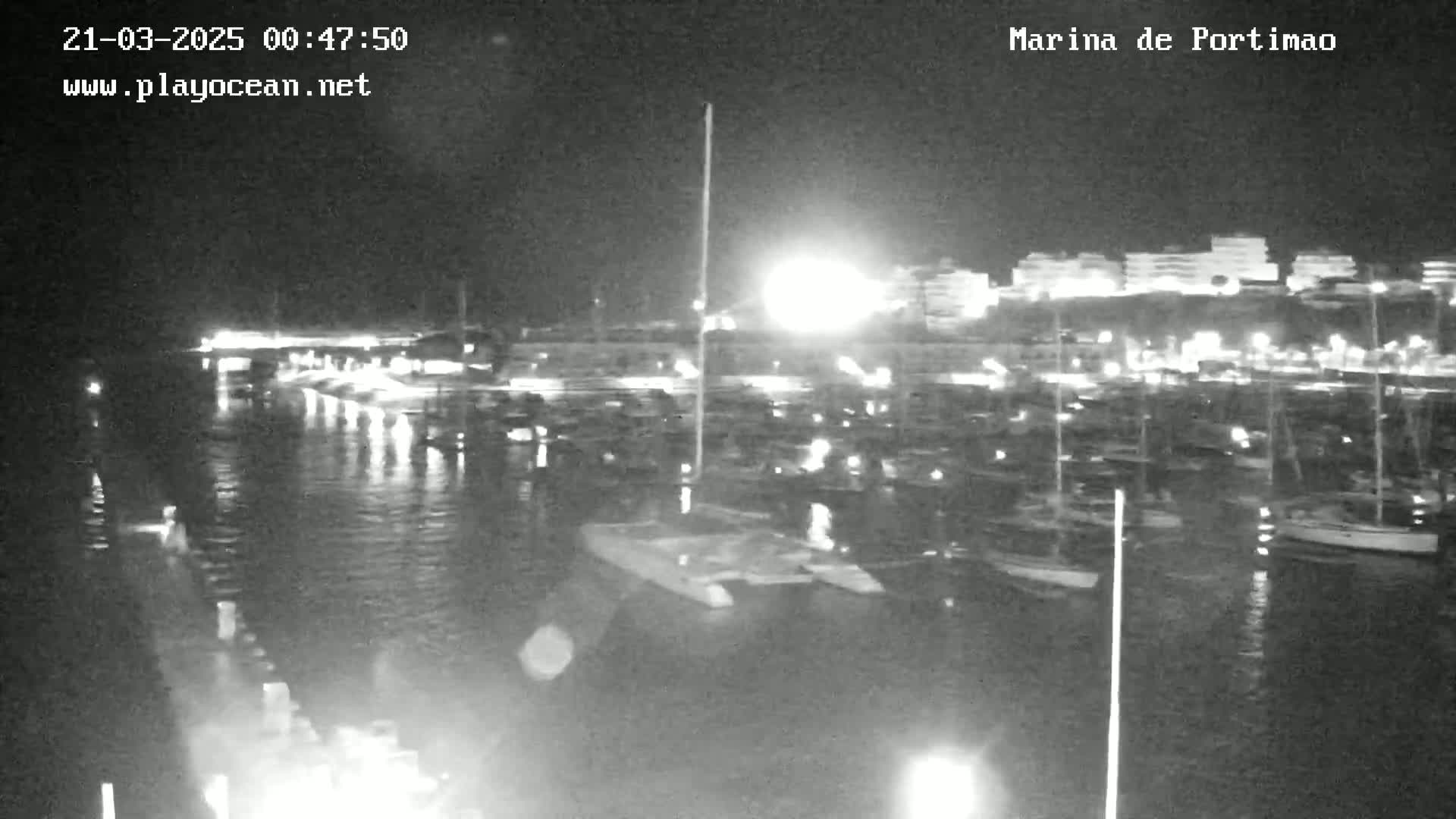 A nighttime, low-light view of a marina filled with boats, with city lights visible in the background.