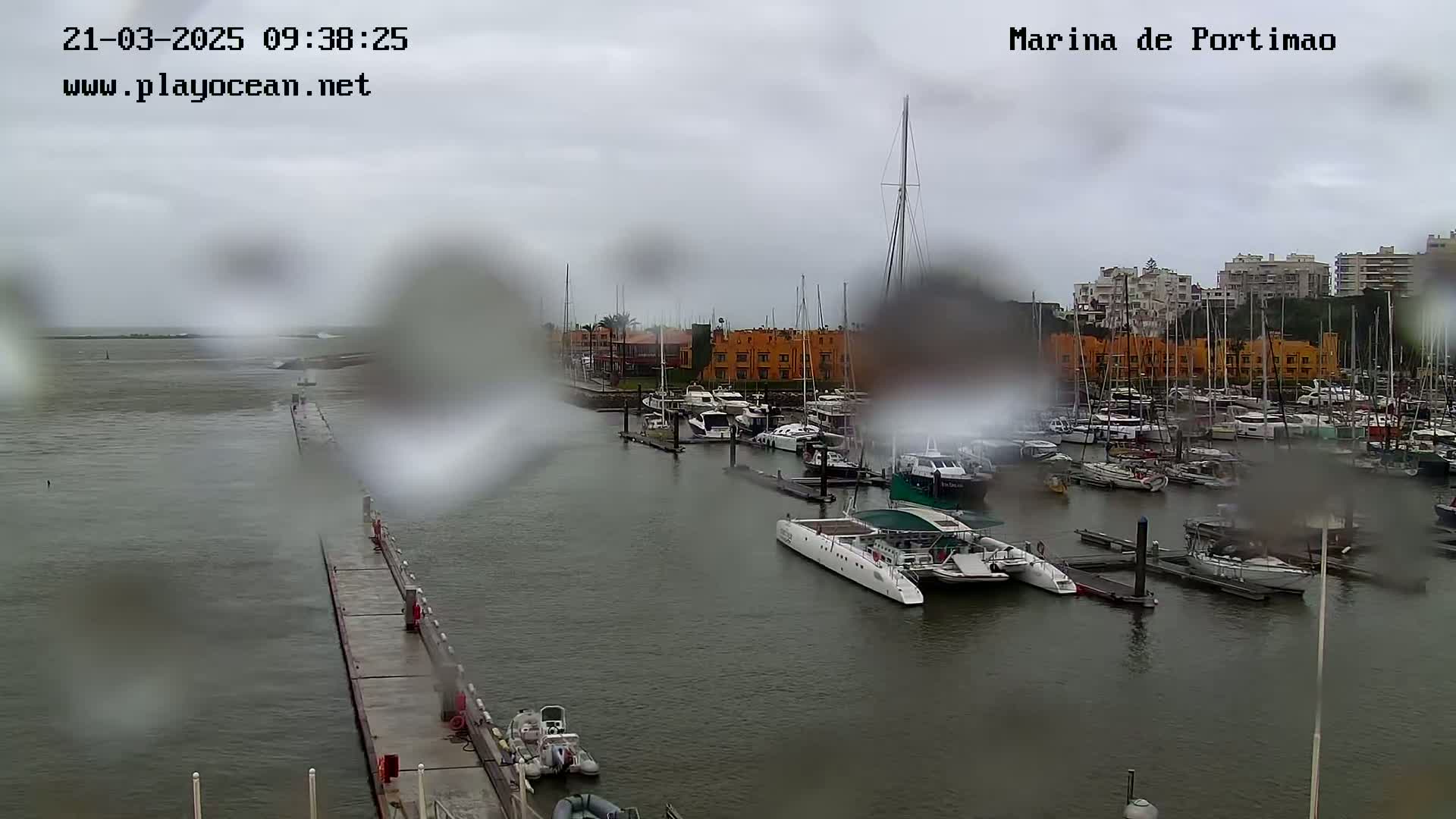 A rain-covered view of a marina filled with boats, with a long pier extending into the water.