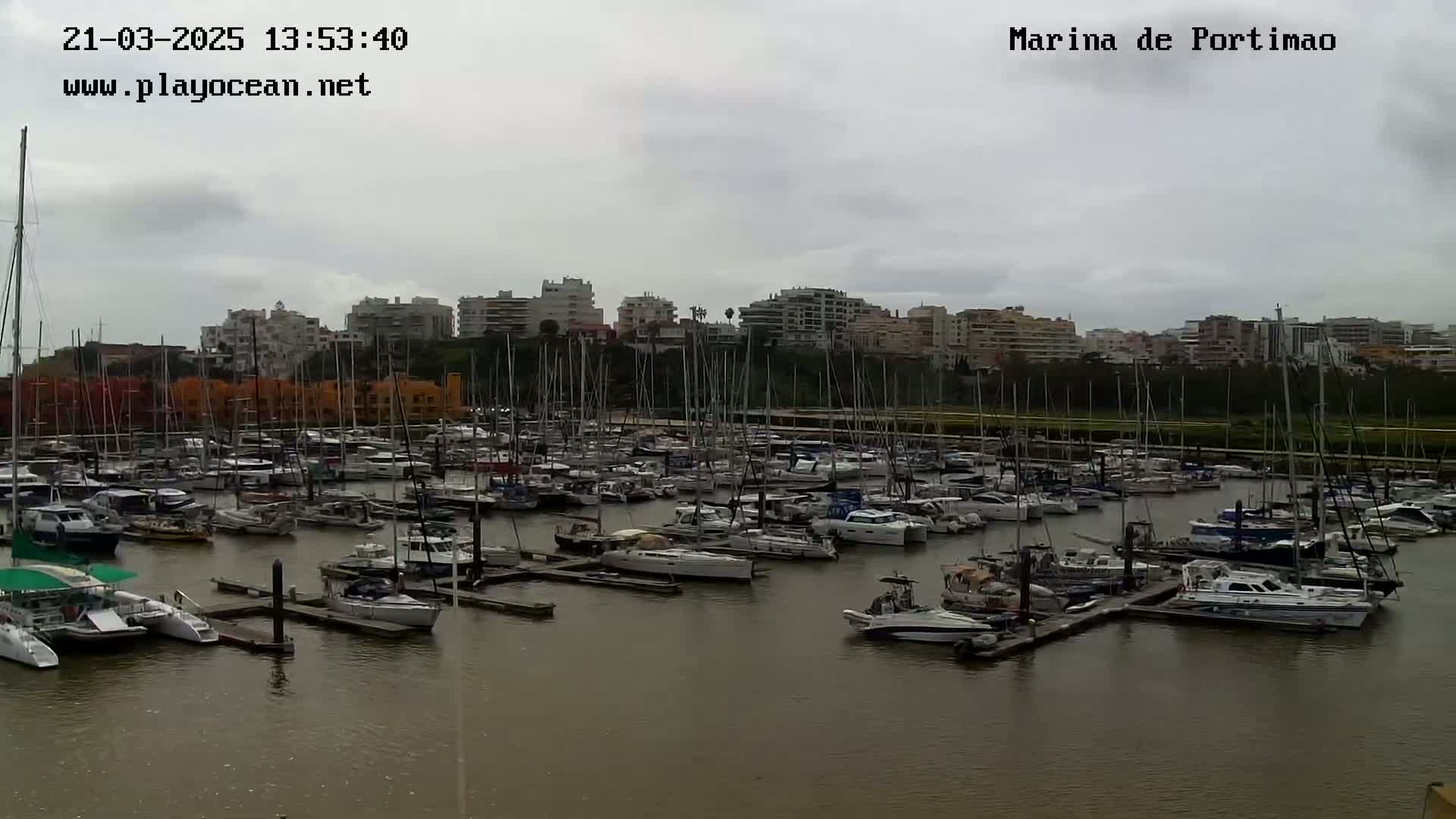 A marina filled with numerous sailboats and motorboats is shown under an overcast sky.