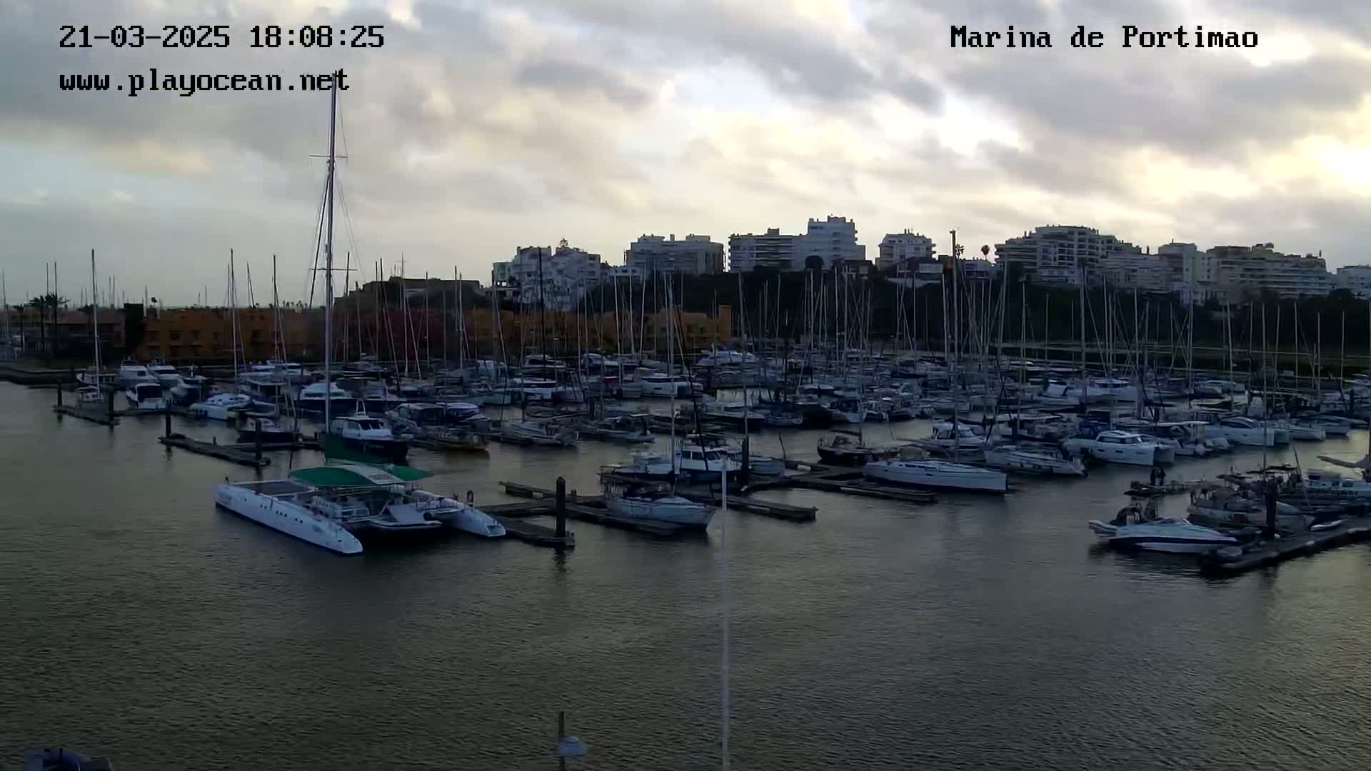 A marina filled with numerous sailboats and motorboats is situated under a cloudy sky at dusk.