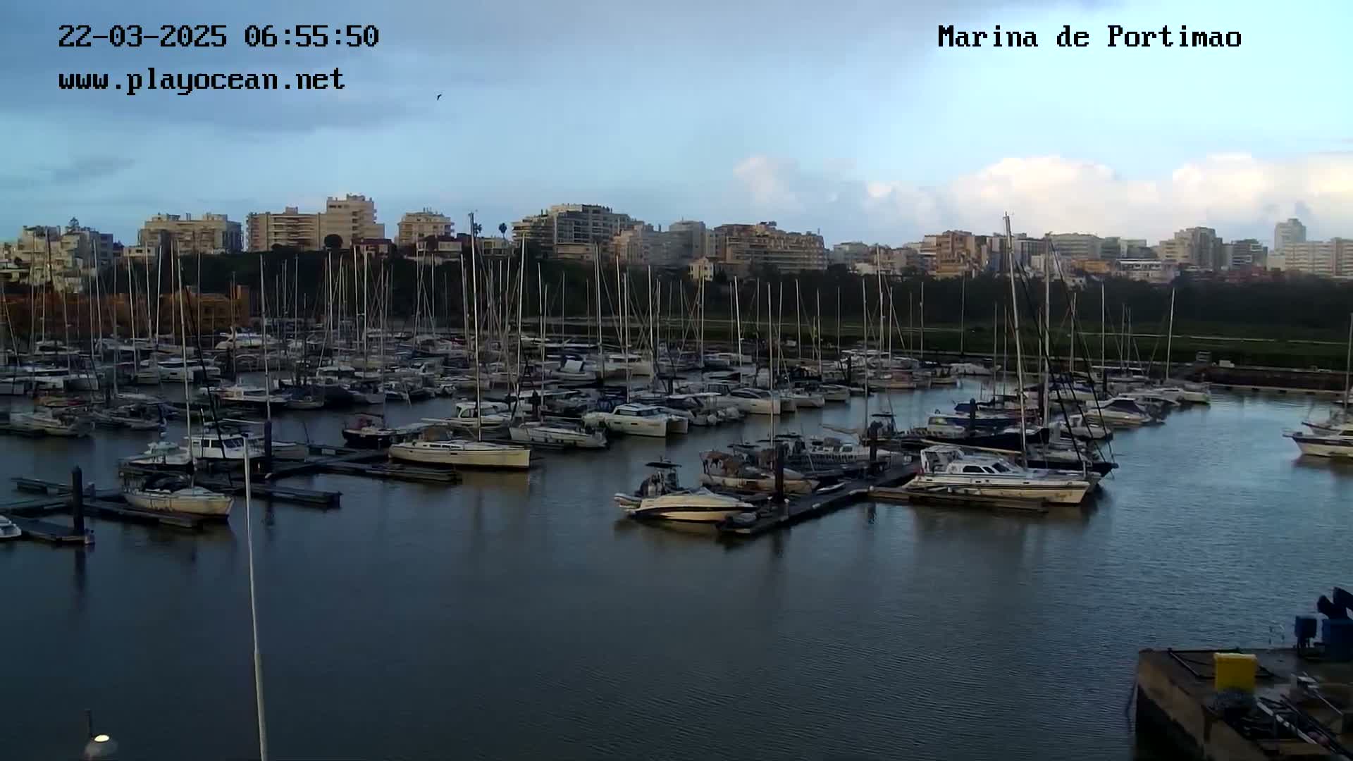 A marina filled with numerous sailboats and motorboats is seen under a partly cloudy sky.