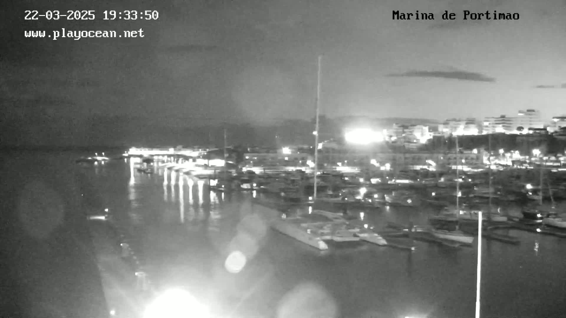 A nighttime, low-light view of a marina filled with boats, with calm water reflecting the lights of the harbor and surrounding buildings.