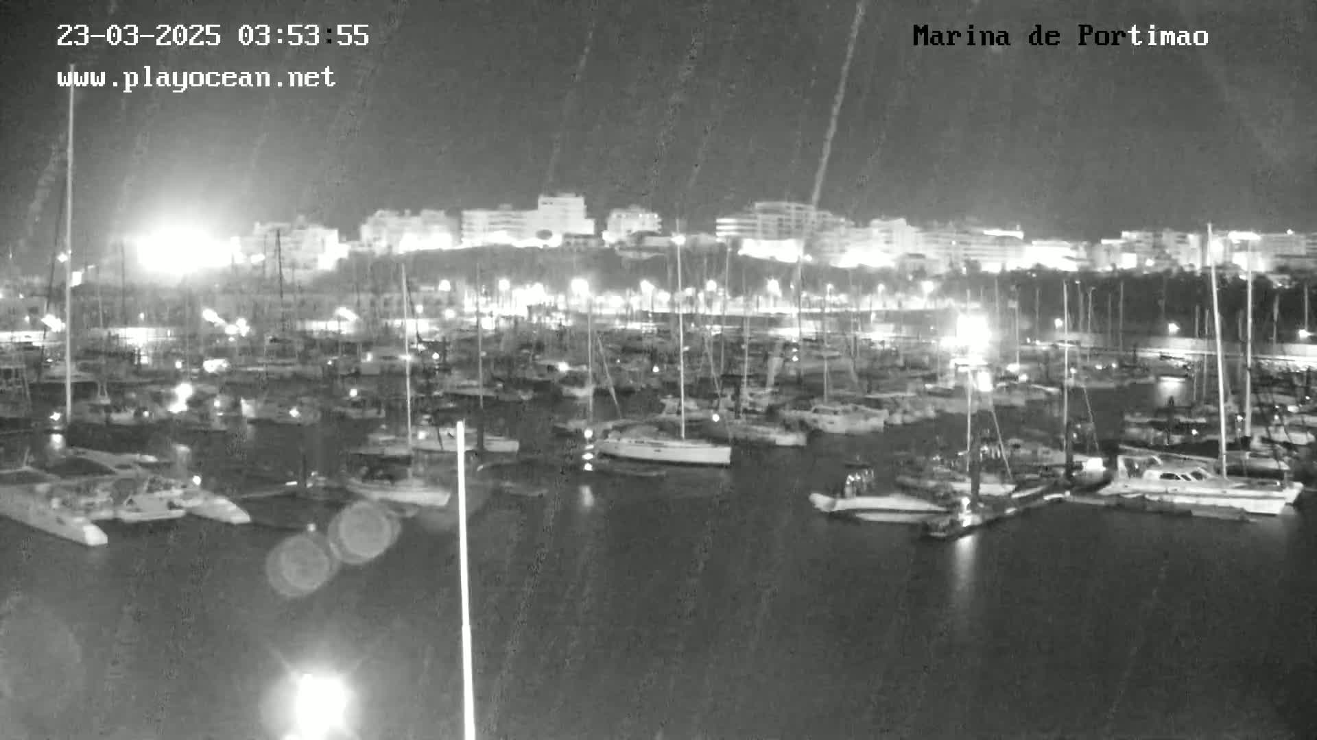 A nighttime view of a marina filled with numerous boats and yachts under a rain shower, with illuminated buildings visible in the background.