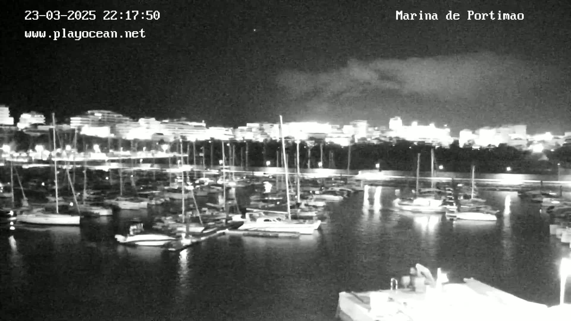 A nighttime long shot of a marina filled with sailboats and yachts, under a partly cloudy sky.