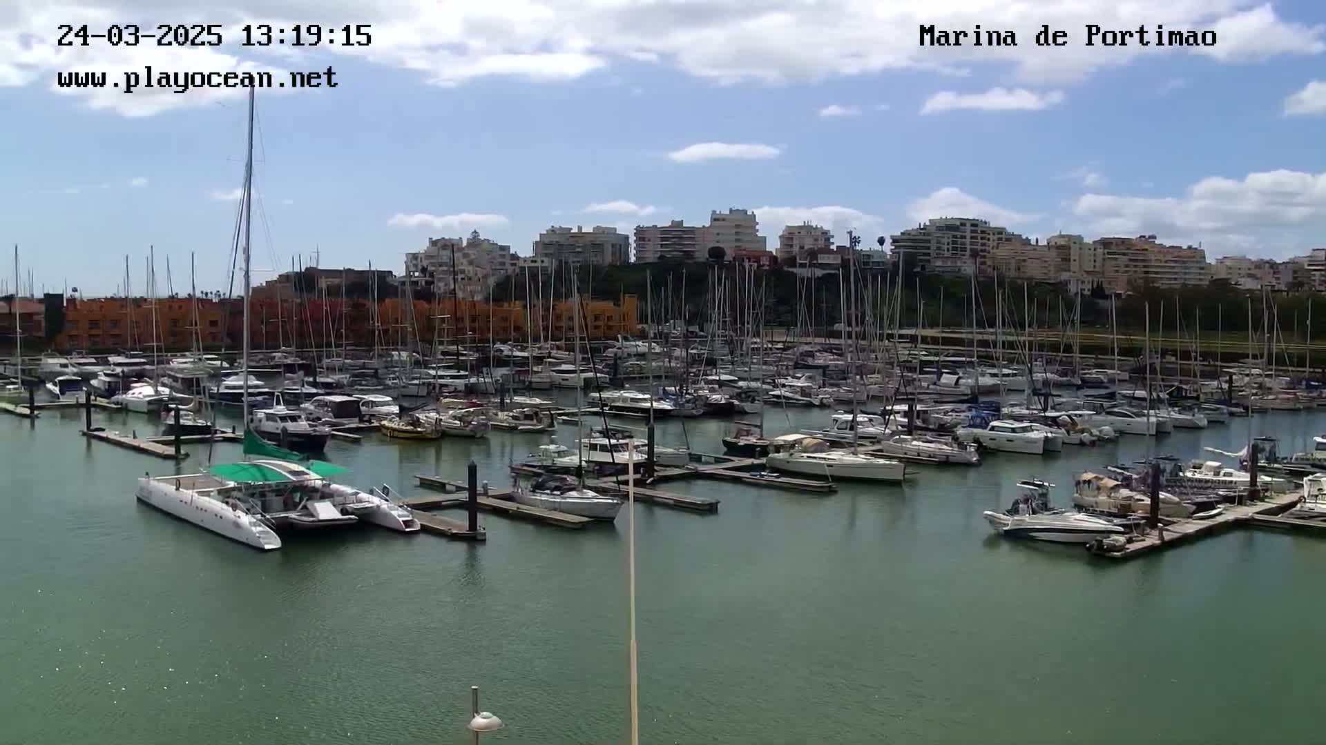 A marina filled with numerous sailboats and motorboats is seen on a partly cloudy day.