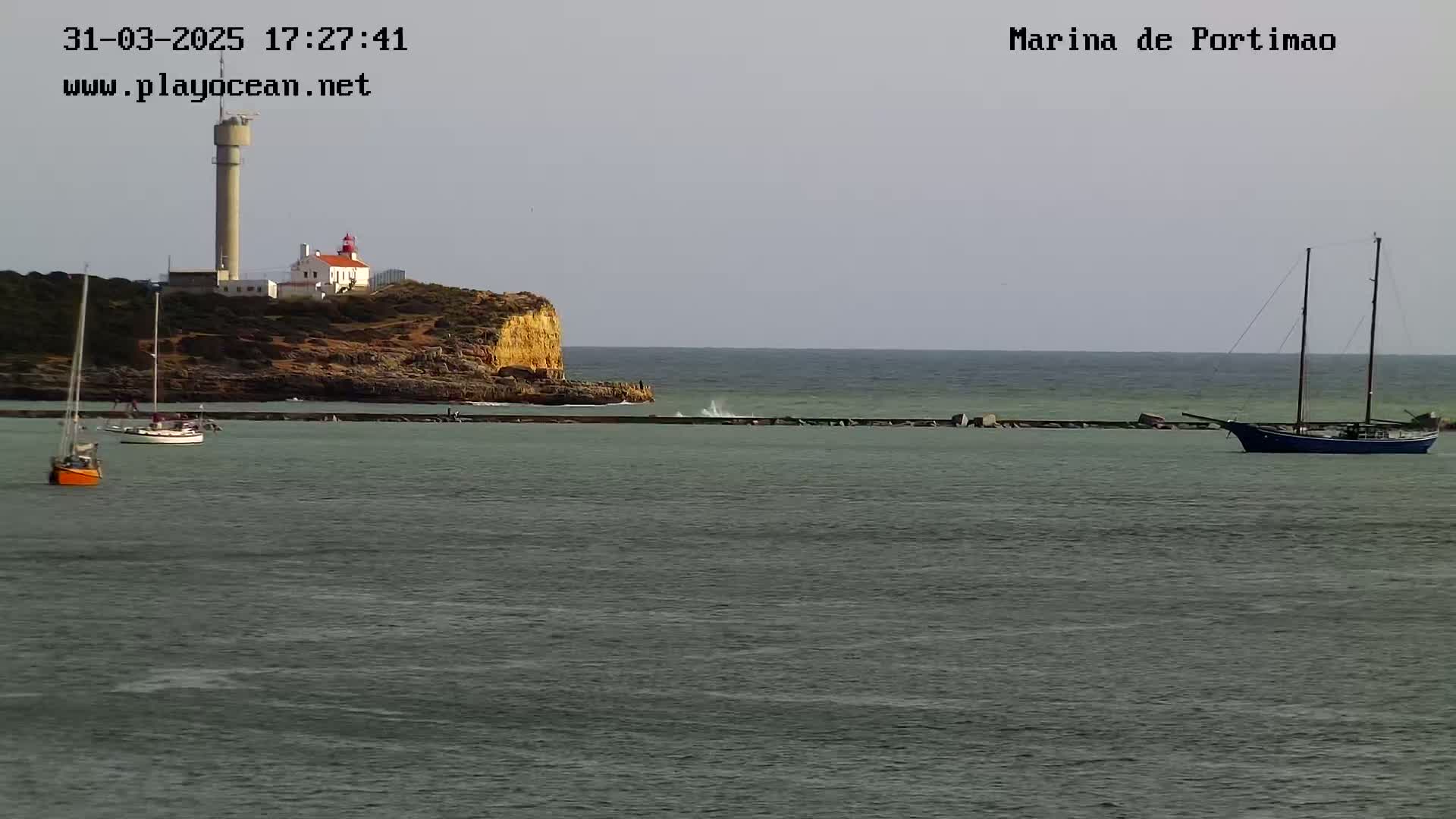A calm sea with several sailboats near a rocky cliff topped with a lighthouse and a tall tower under a mostly clear sky.