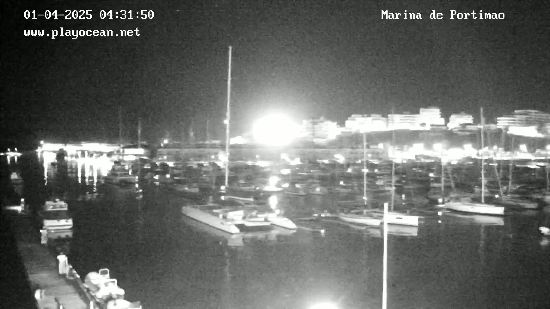 A nighttime, low-light view of a marina filled with numerous boats and yachts, illuminated by lights from the vessels and a nearby shoreline.
