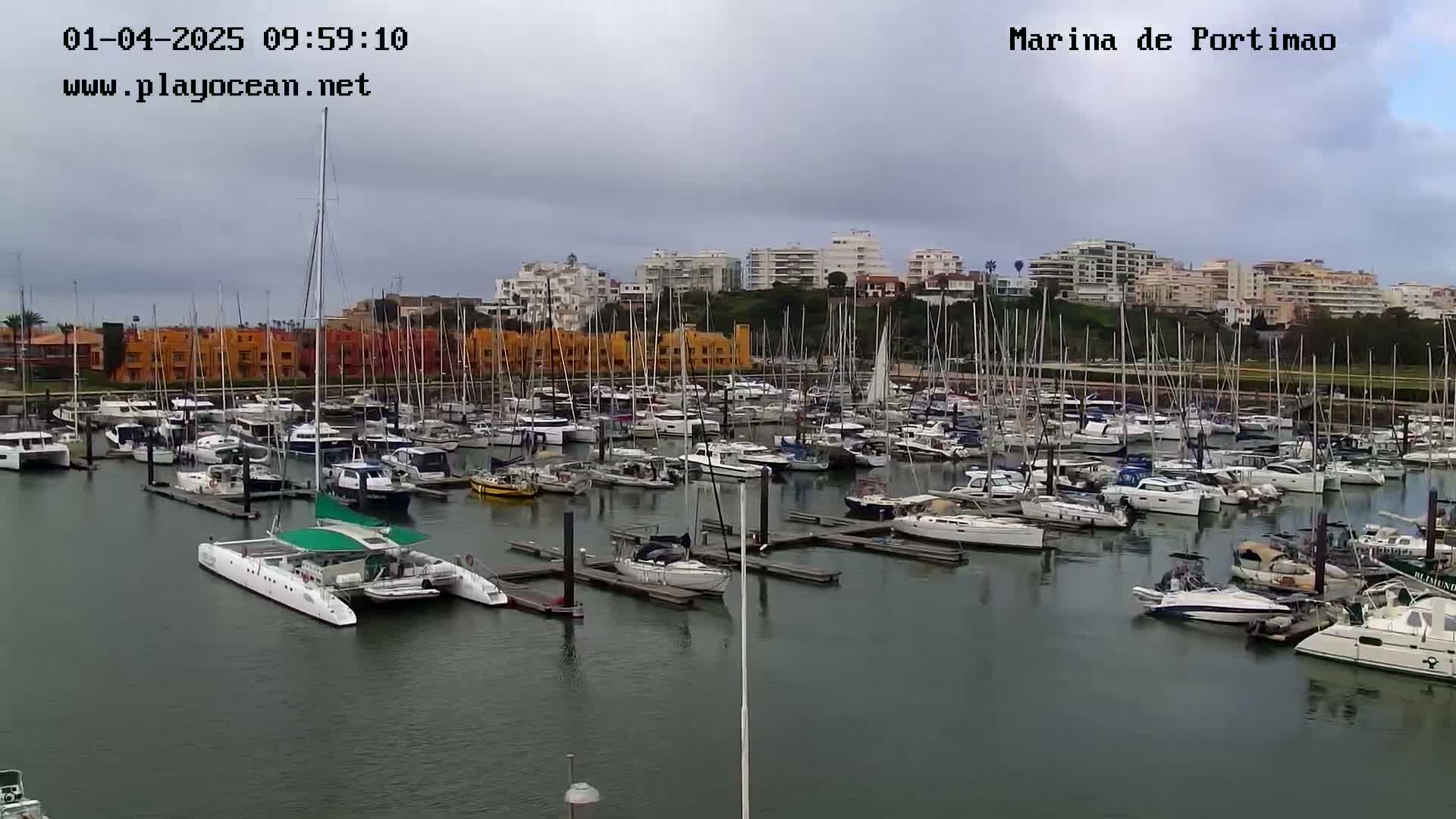 A marina filled with numerous sailboats and motorboats is shown under an overcast sky.