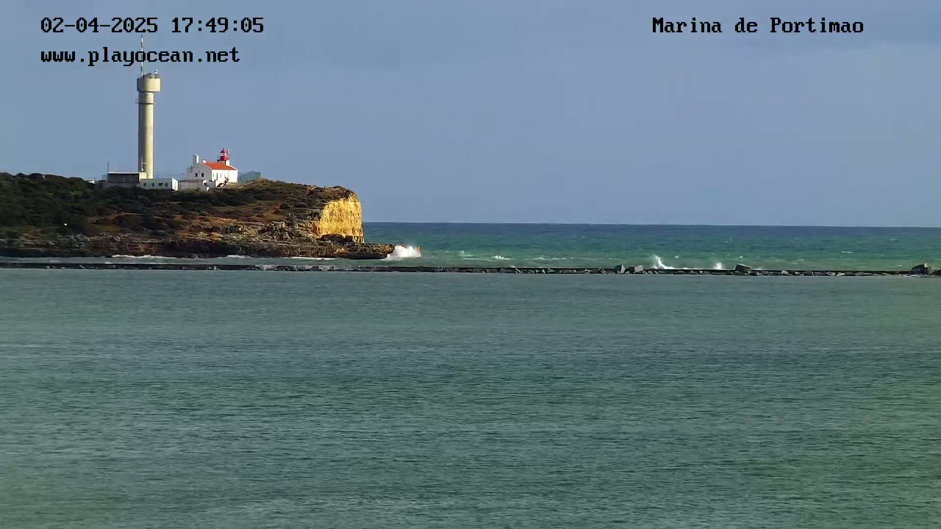 A lighthouse sits atop a rocky cliff overlooking a calm sea with small waves under a clear sky.