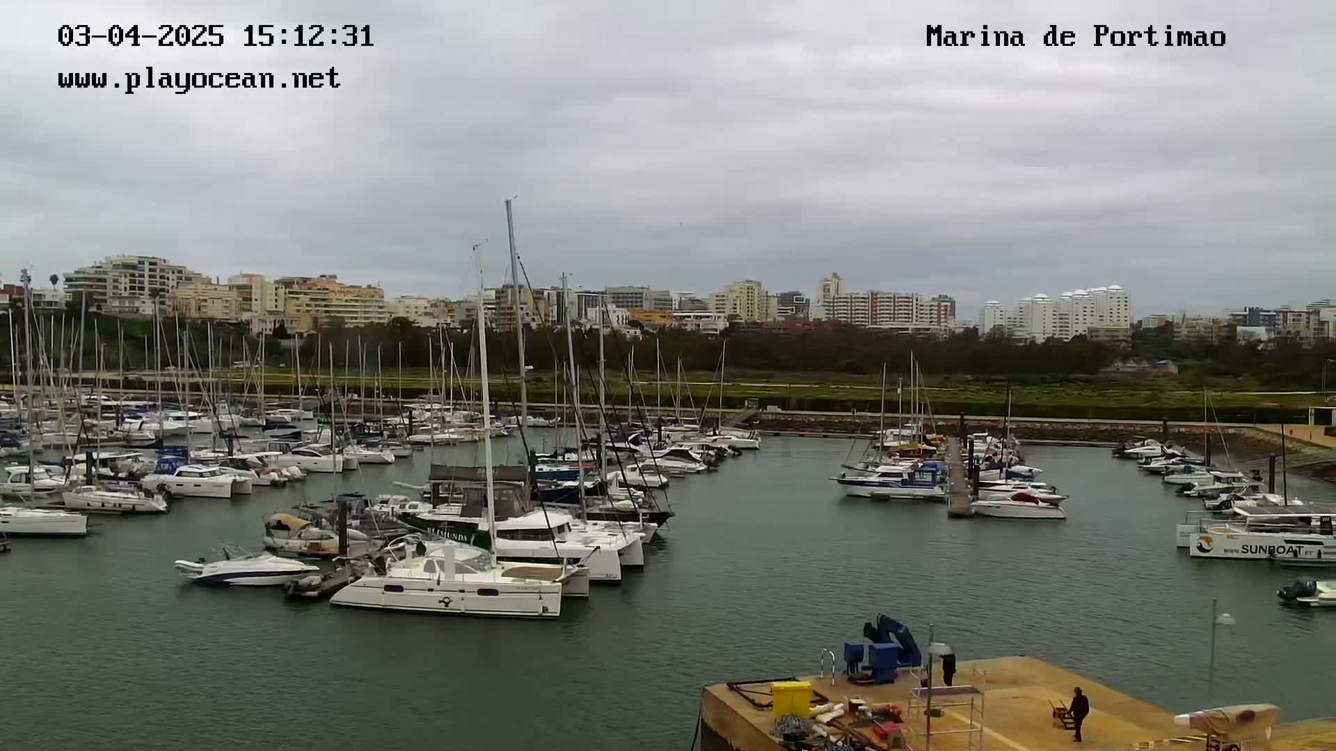 A marina filled with numerous sailboats and motorboats is situated under an overcast sky, with a cityscape visible in the background.