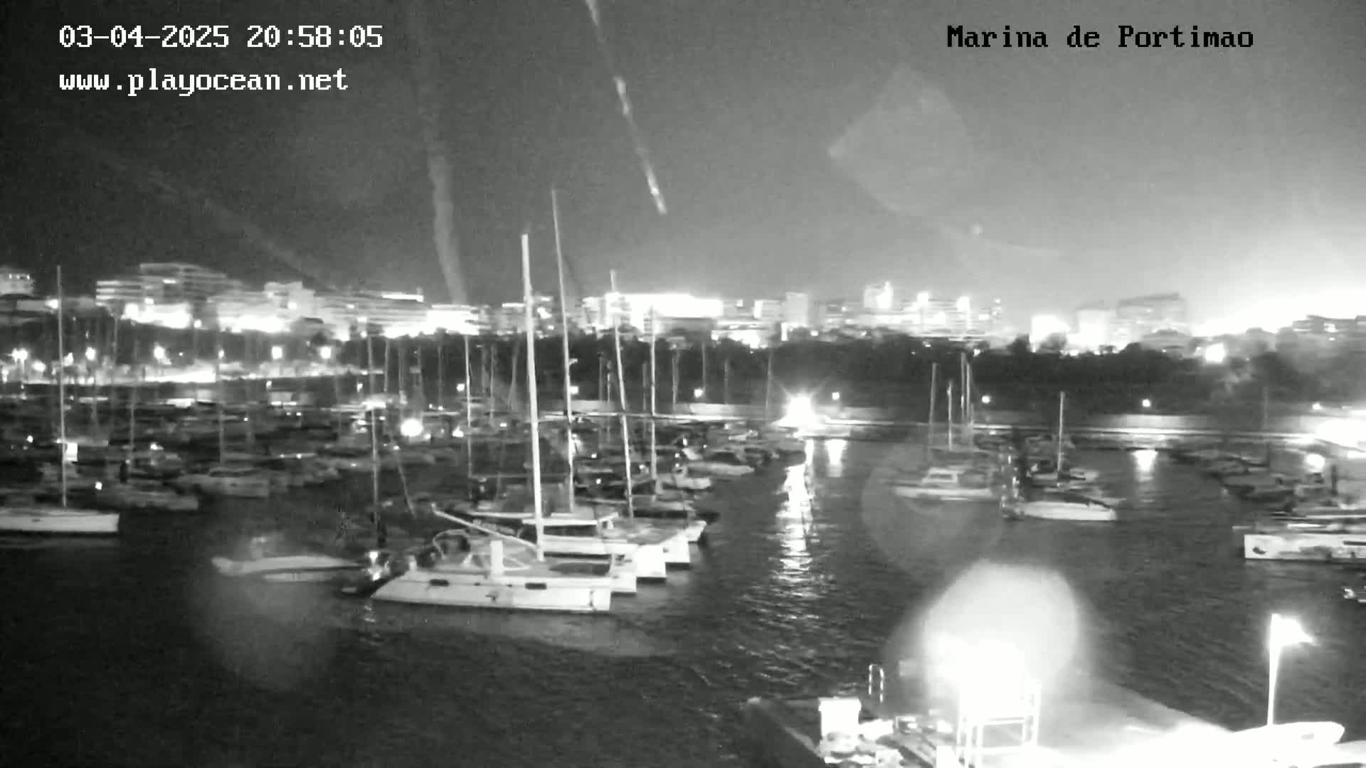 A nighttime view of a marina filled with sailboats, with city lights visible in the background under a partly cloudy sky.