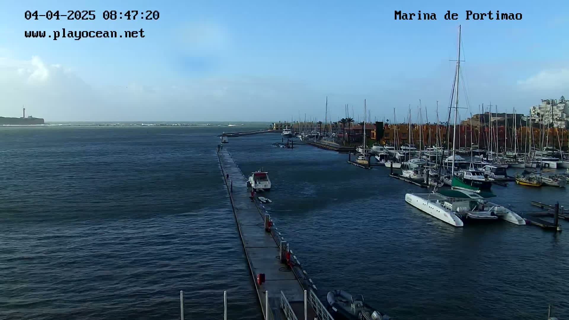 A marina filled with boats on a calm, clear day, with a long pier extending into the water toward a distant landmass.