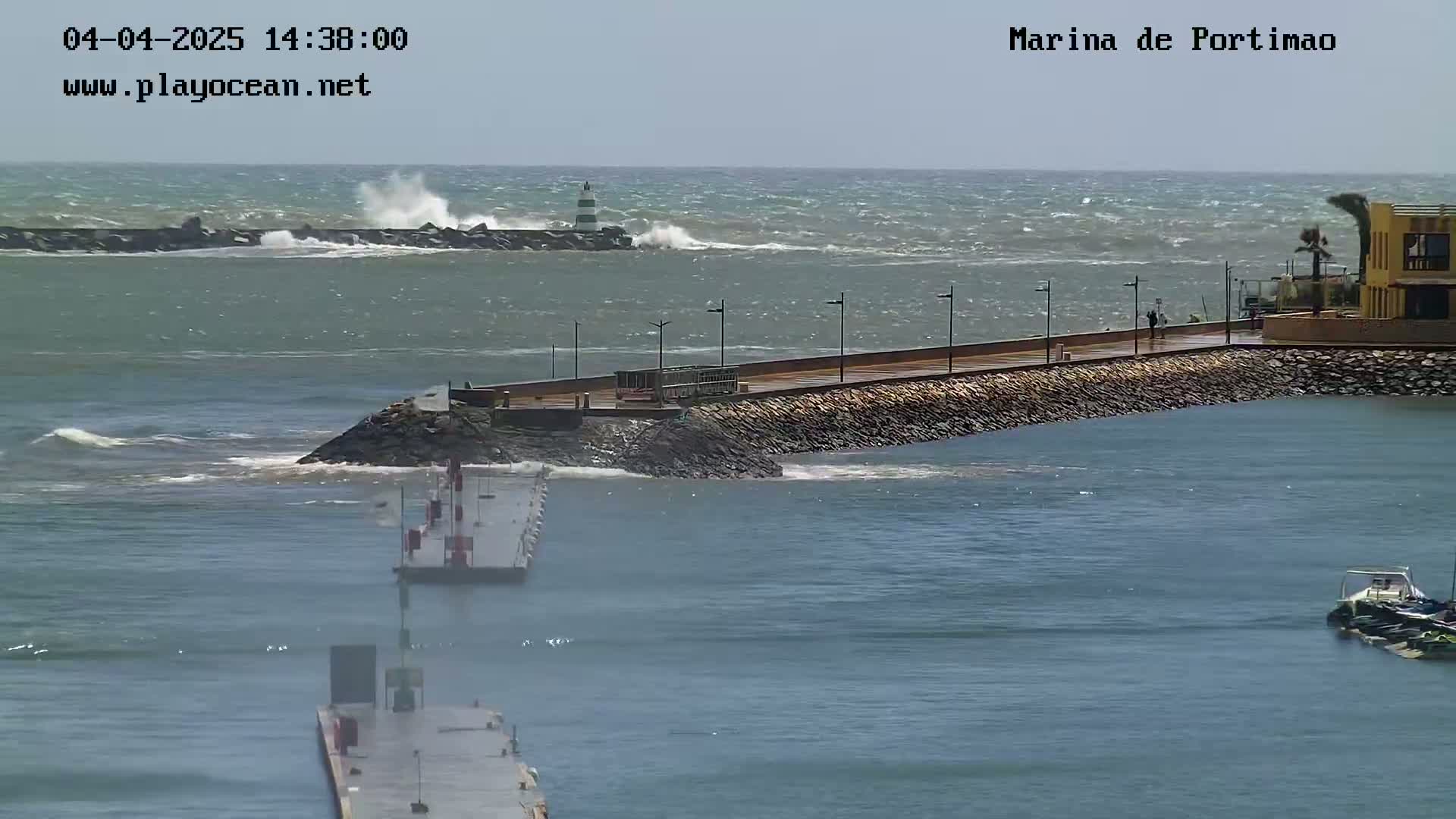 A marina with a walkway and a lighthouse is partially protected by a breakwater from a windy, wavy ocean.
