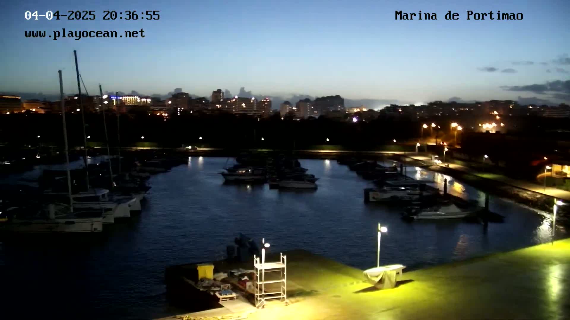 A nighttime view of a marina filled with boats, with a city skyline illuminated in the distance under a twilight sky.