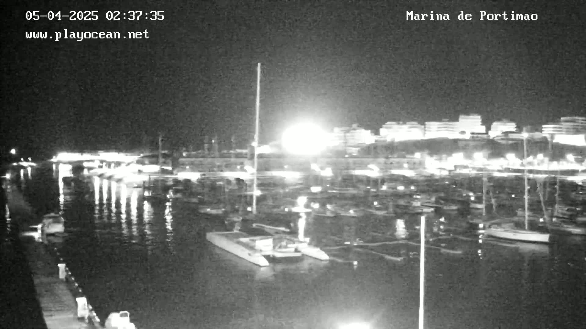 A nighttime view of a marina filled with boats, illuminated by lights from the buildings and boats on the water.
