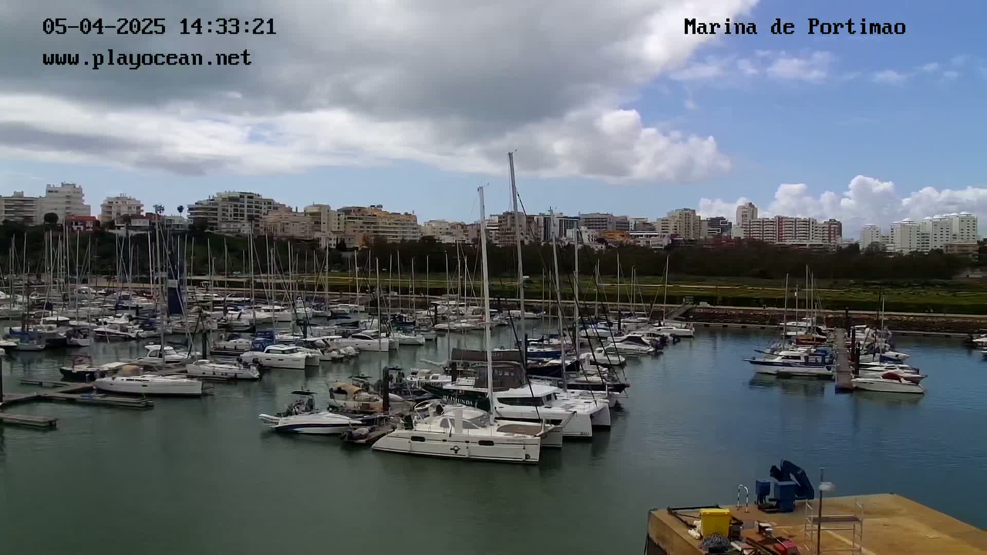 A marina filled with numerous sailboats and motorboats is situated in front of a shoreline with buildings under a partly cloudy sky.