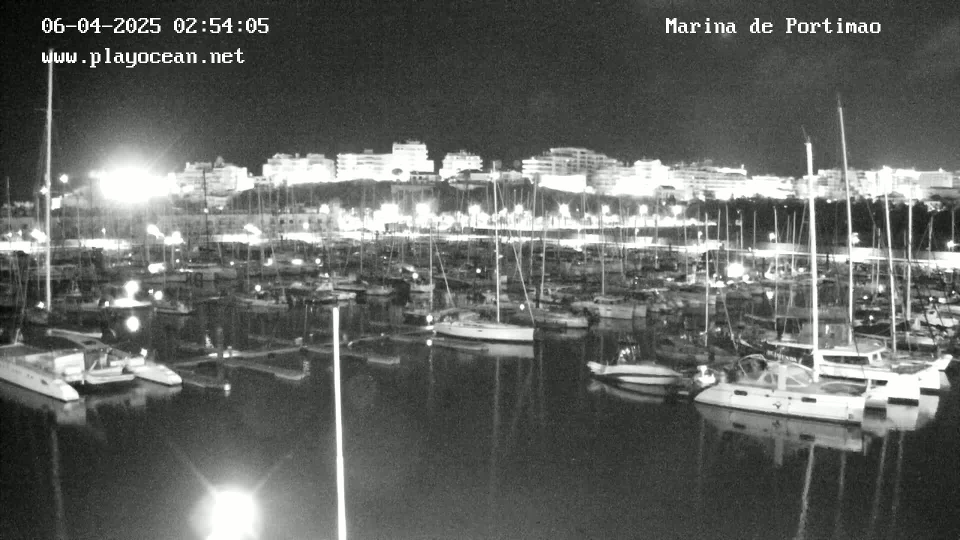 A nighttime, black and white view of a marina filled with many boats and yachts, illuminated by lights on the shore and boats.