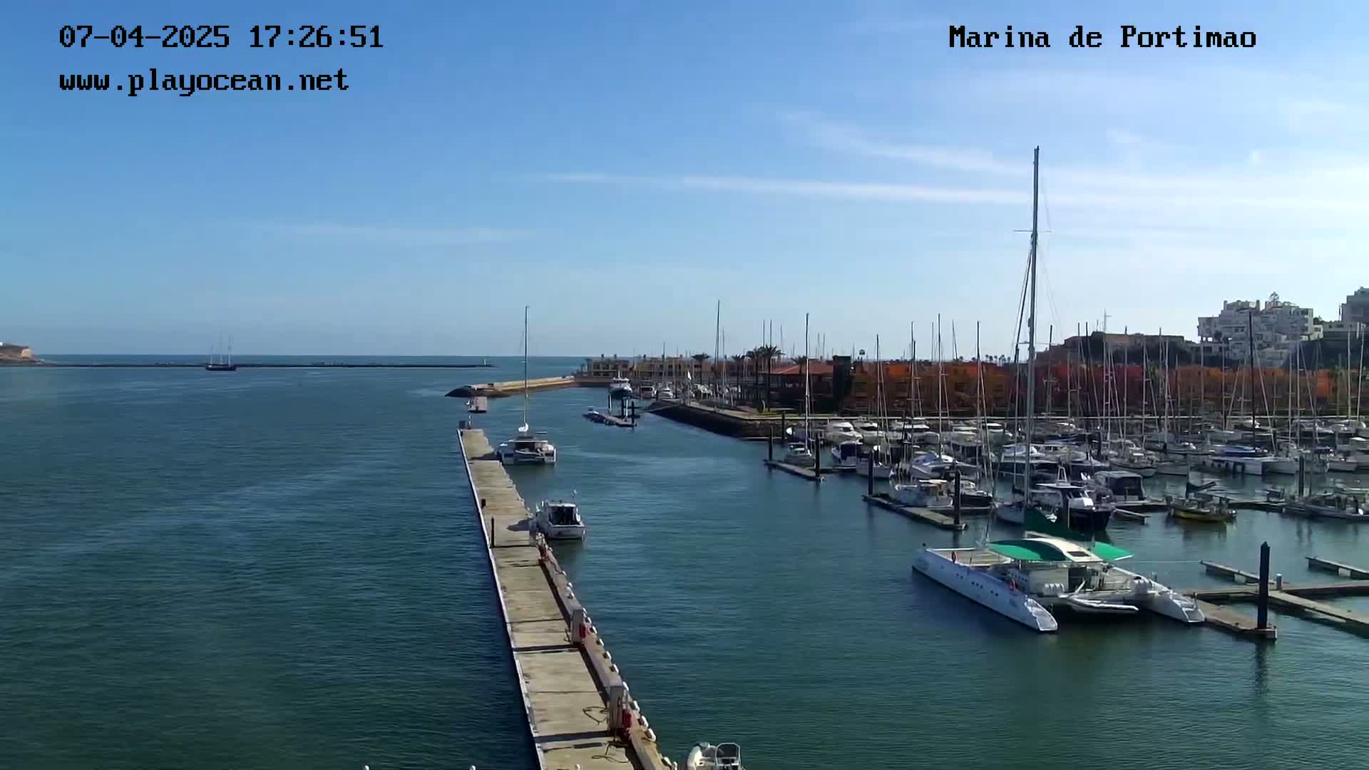 A marina filled with numerous boats and yachts on a sunny day, with calm water and a long dock extending into the harbor.