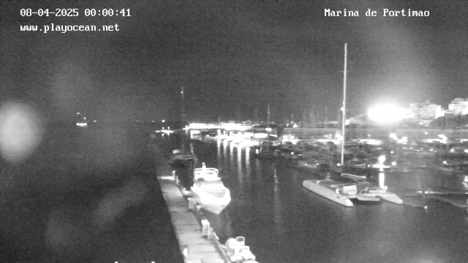A nighttime, low-light view of a marina filled with boats and yachts, with some lights visible on land in the background.
