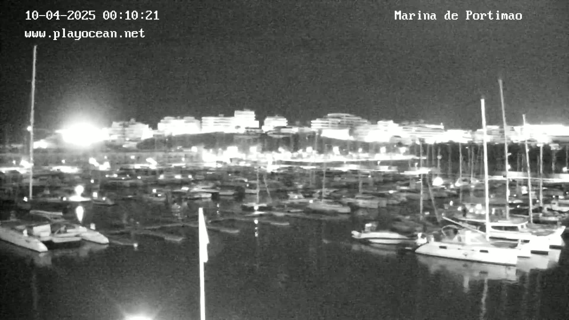 A nighttime, low-light view of a marina filled with numerous boats, with a town's lights visible in the background.