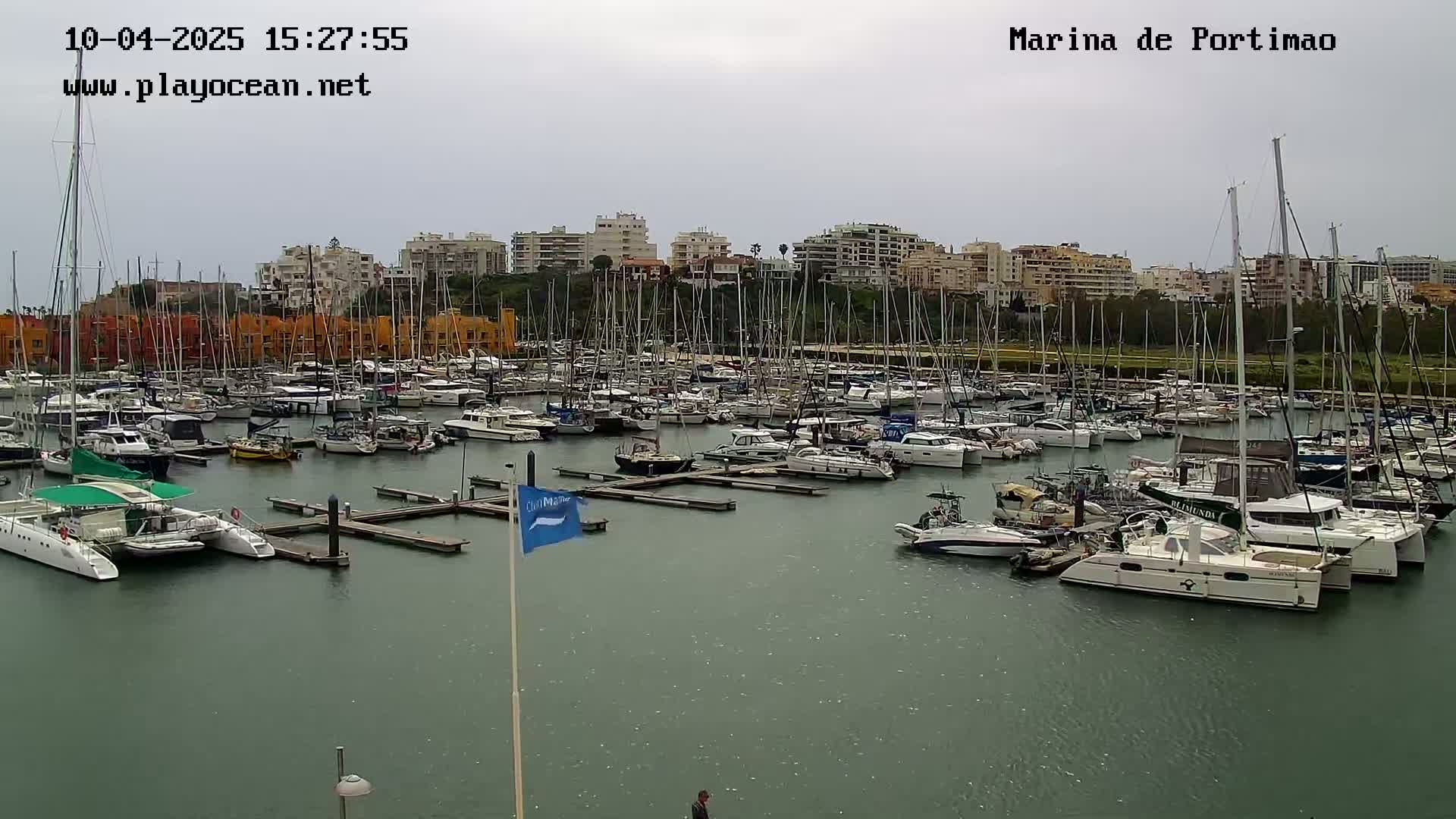 A marina filled with numerous sailboats and yachts is visible under an overcast sky.