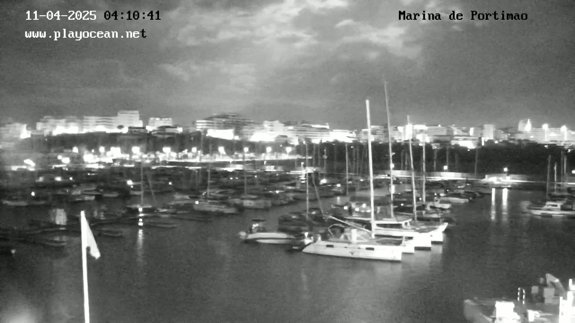 A nighttime view of a marina filled with boats, under a partly cloudy sky, with a city skyline illuminated in the background.