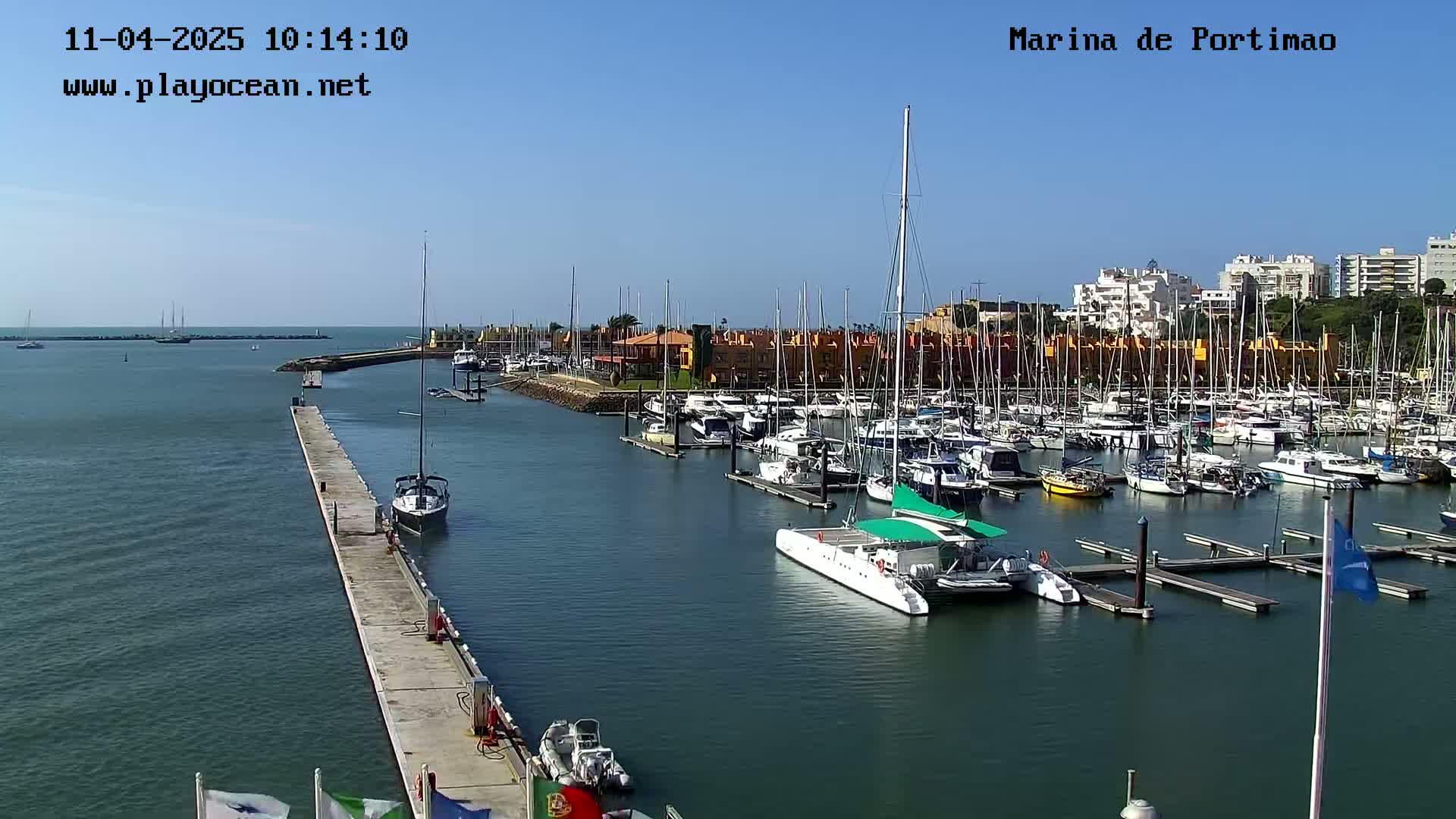 A marina filled with numerous boats and yachts is shown on a sunny day, with a long pier extending into the calm water.