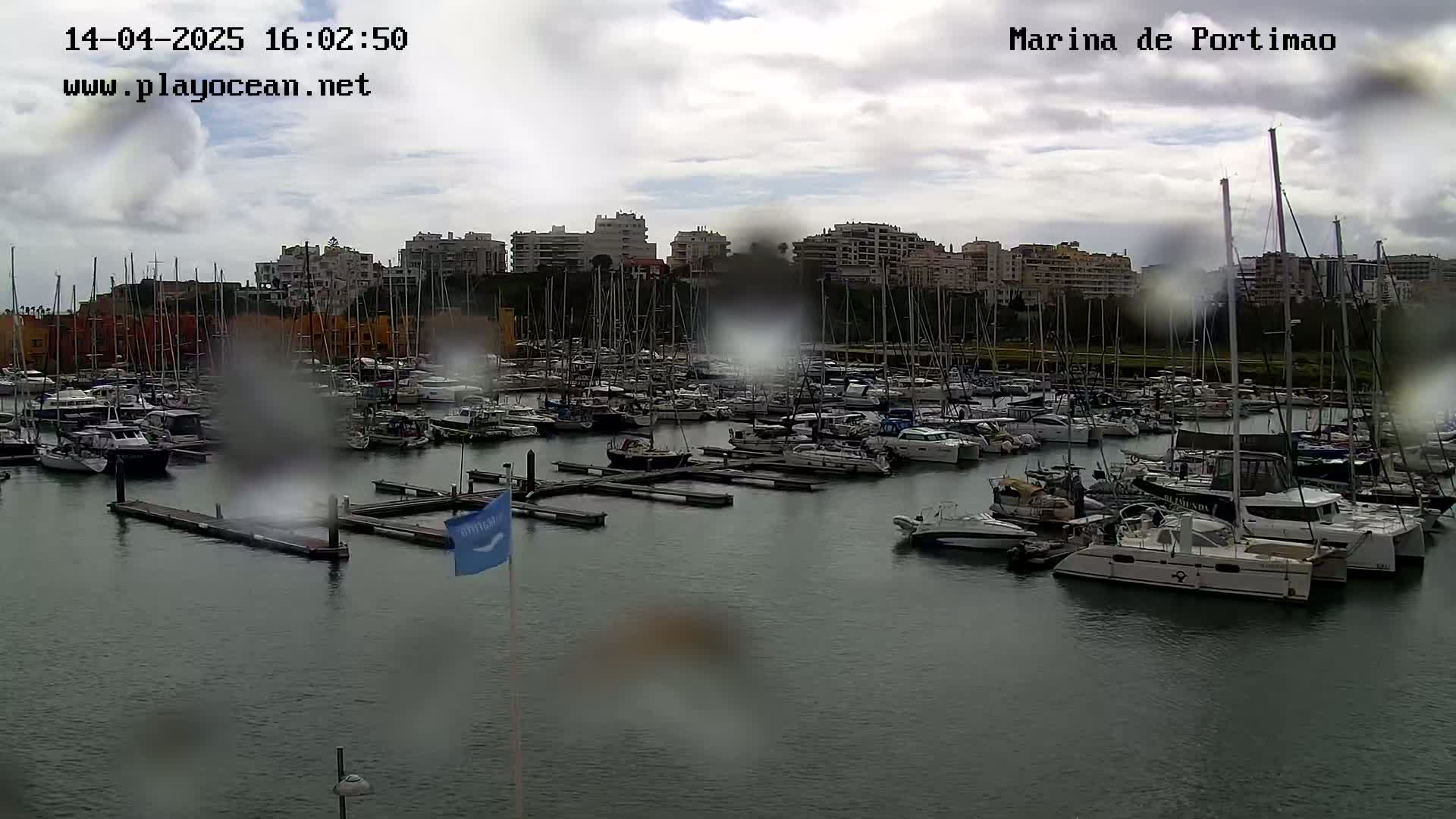 A marina filled with numerous boats and yachts is seen under an overcast, rainy sky.
